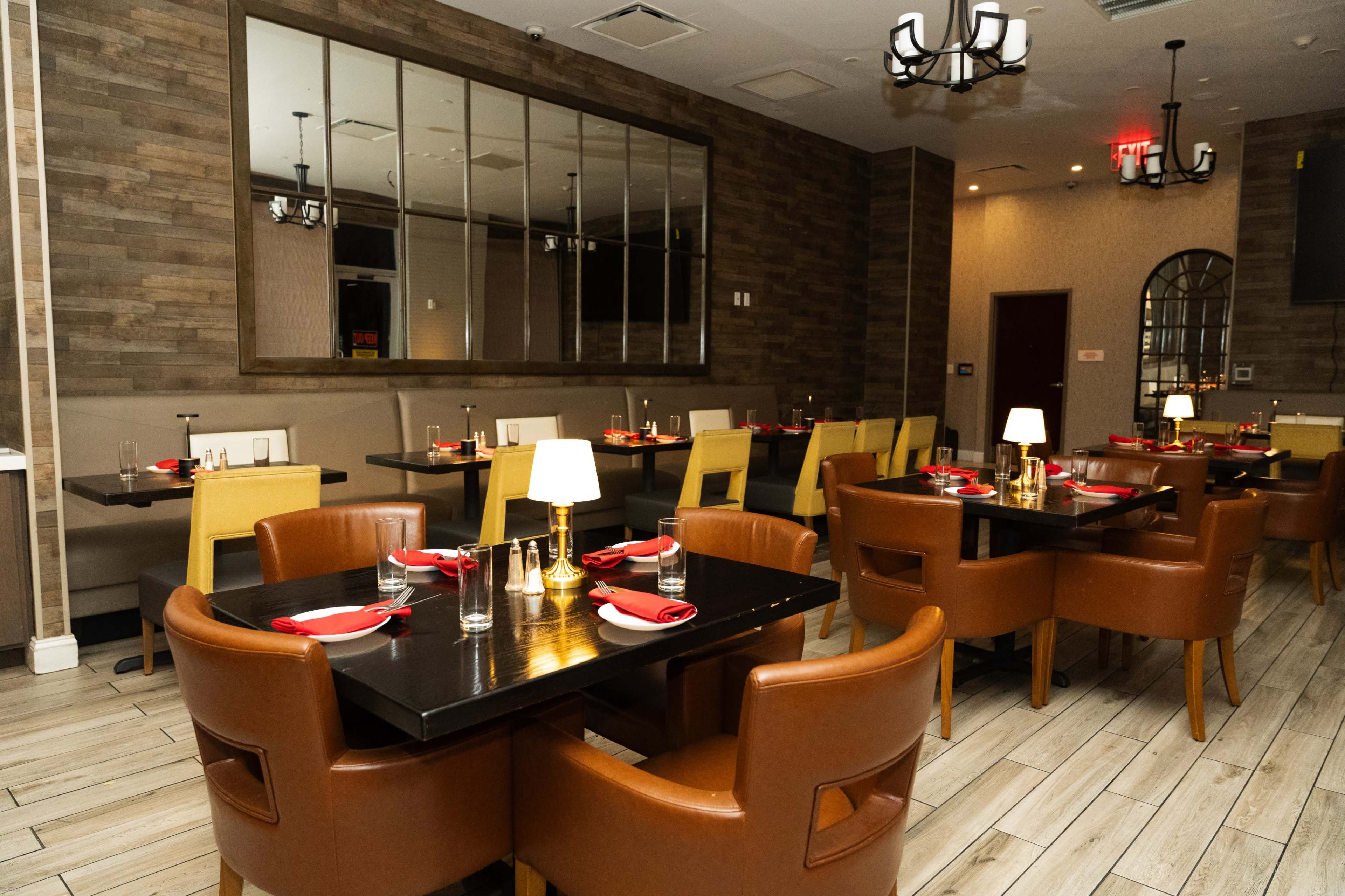 The image shows an upscale restaurant interior featuring dark wooden floors, beige and brown seating, and tables set with red napkins and candles.