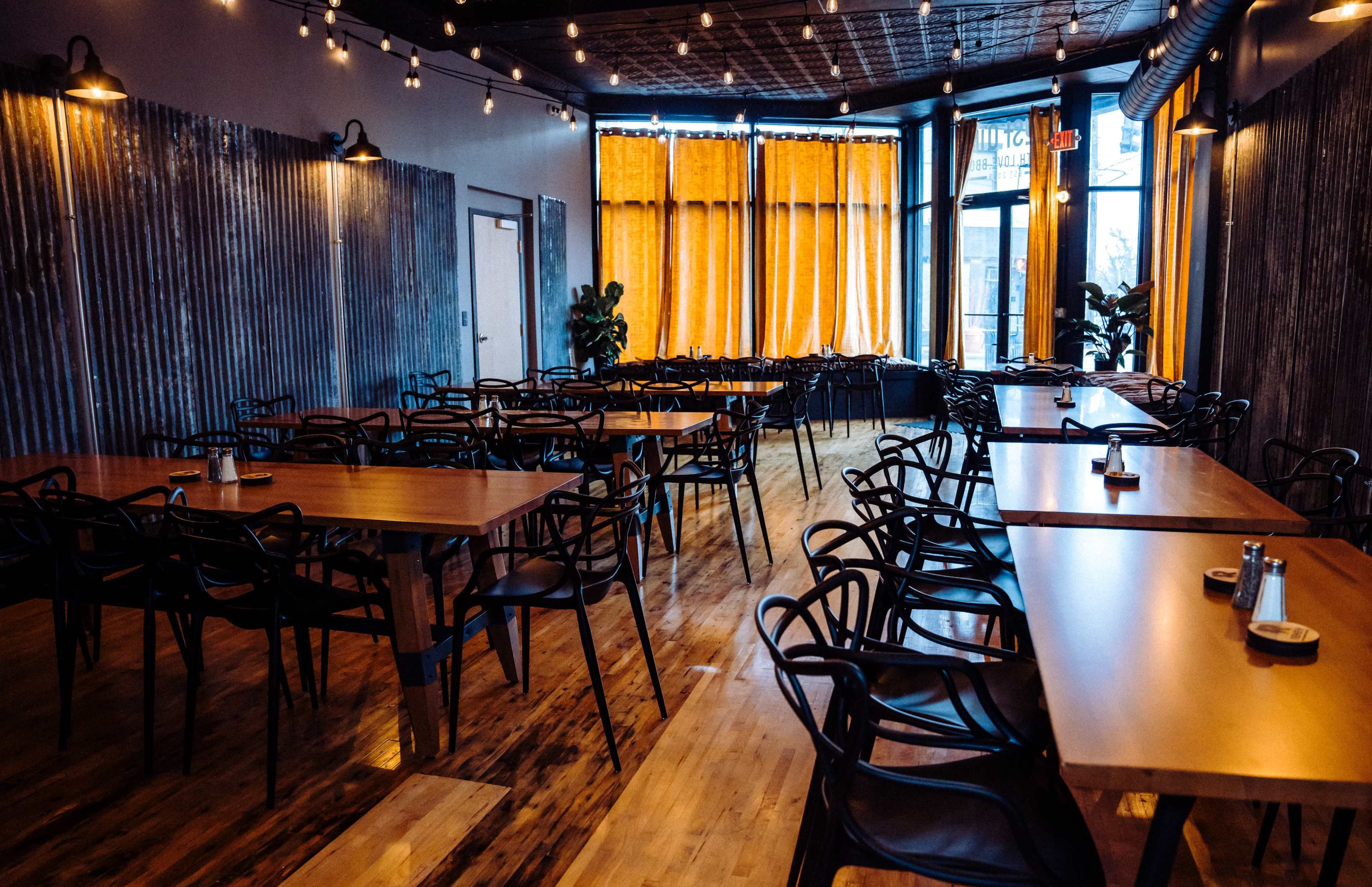 The image shows a spacious restaurant interior with wooden floors, metal accents, and several rows of tables and chairs arranged under warm lighting.