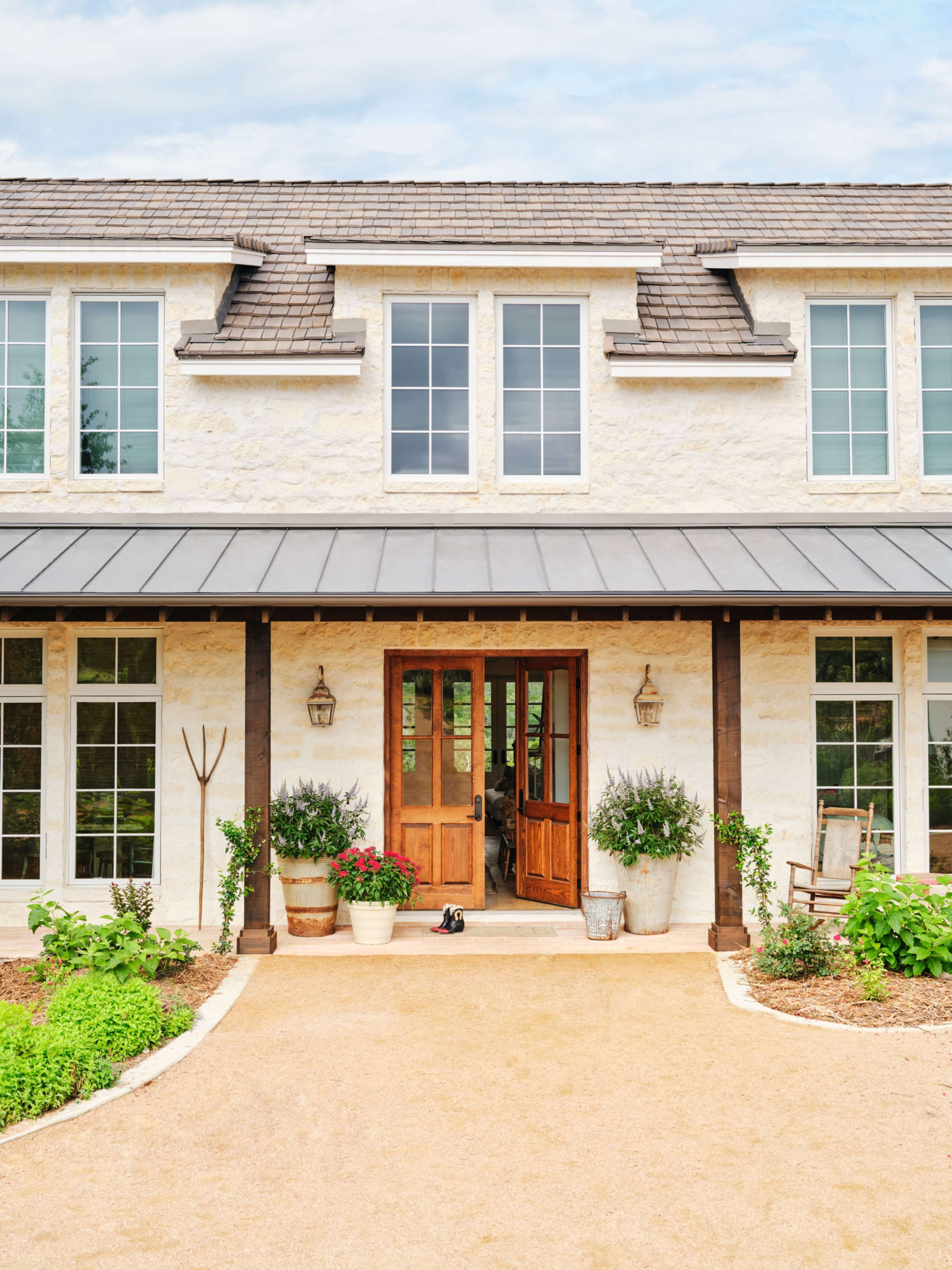 The image shows a modern, two-story house with a light-colored exterior, large windows, and a welcoming front porch surrounded by greenery and potted plants.