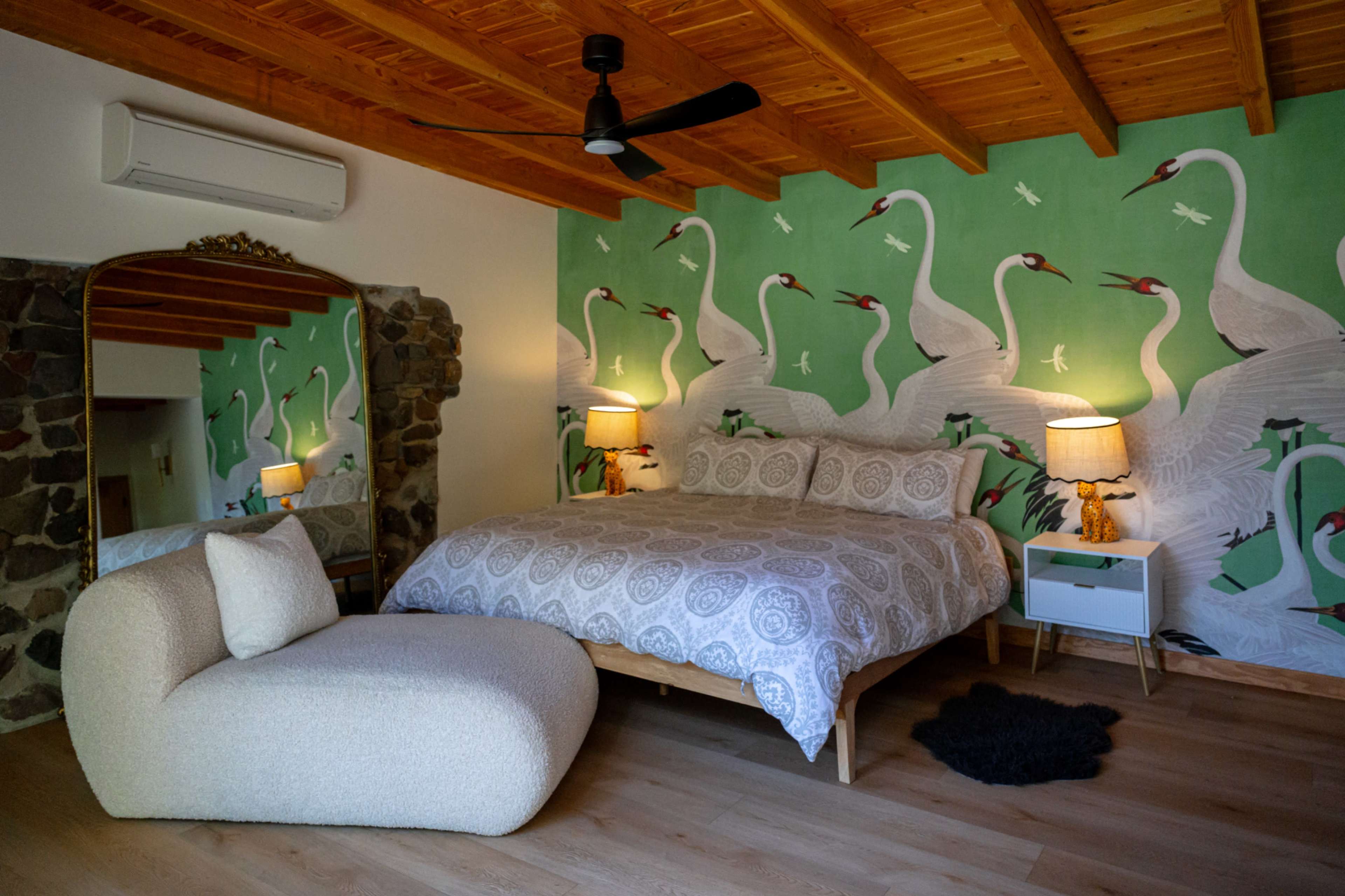 The bedroom features a large bed with a patterned comforter, a cozy lounge chair, a decorative mirror, and walls painted with swan motifs.