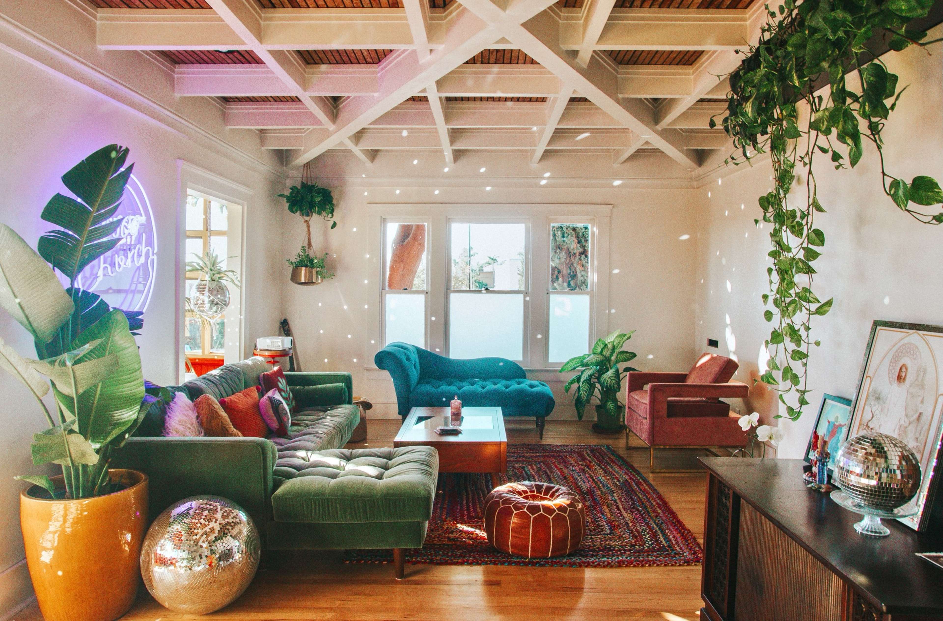 A sunlit living room with a green sofa, a pink chair, a wooden coffee table, plants, and colorful decor on the walls.