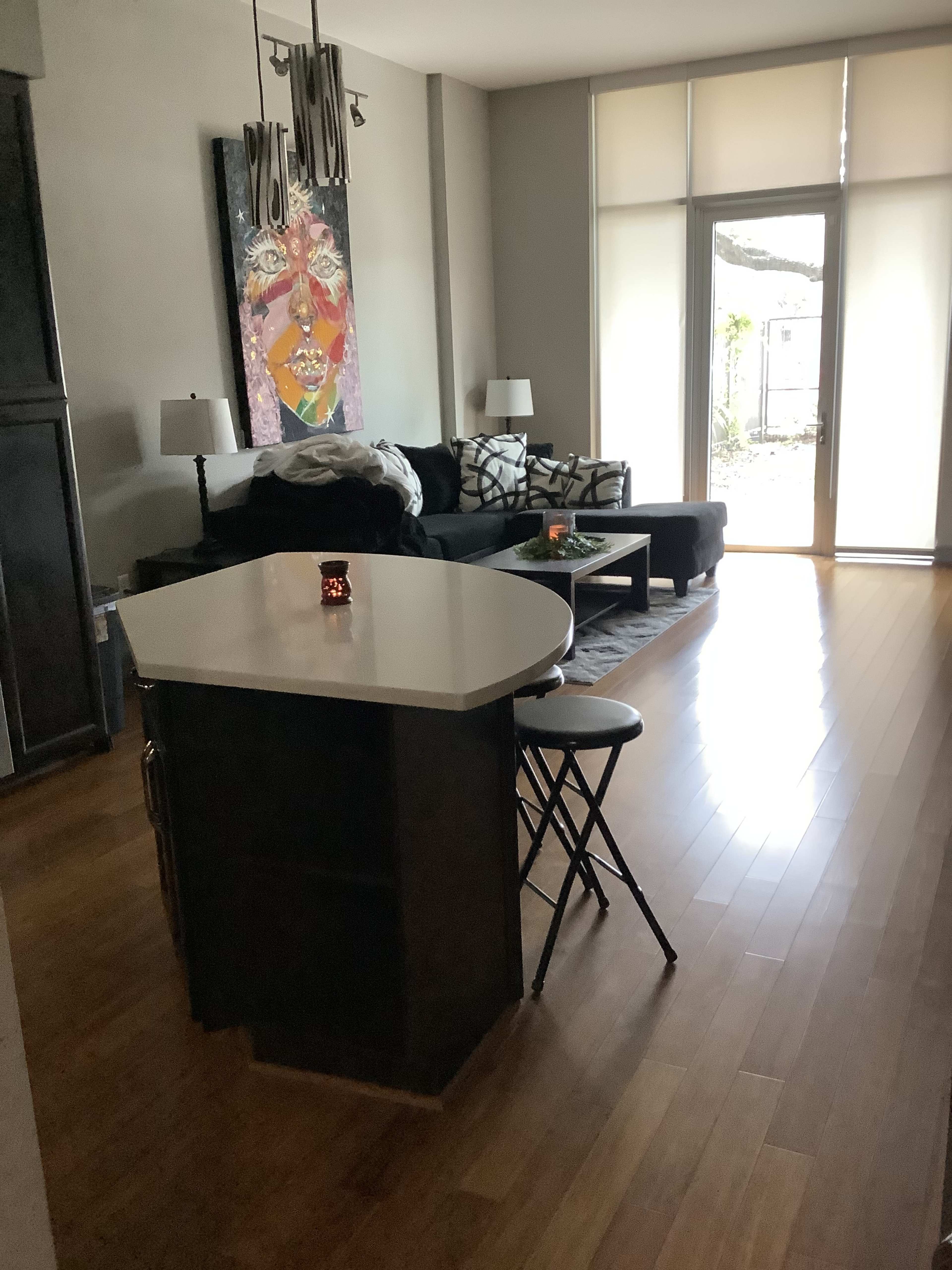 A modern living space features a kitchen island with two stools, a black sofa, artwork on the wall, and large windows allowing natural light in.