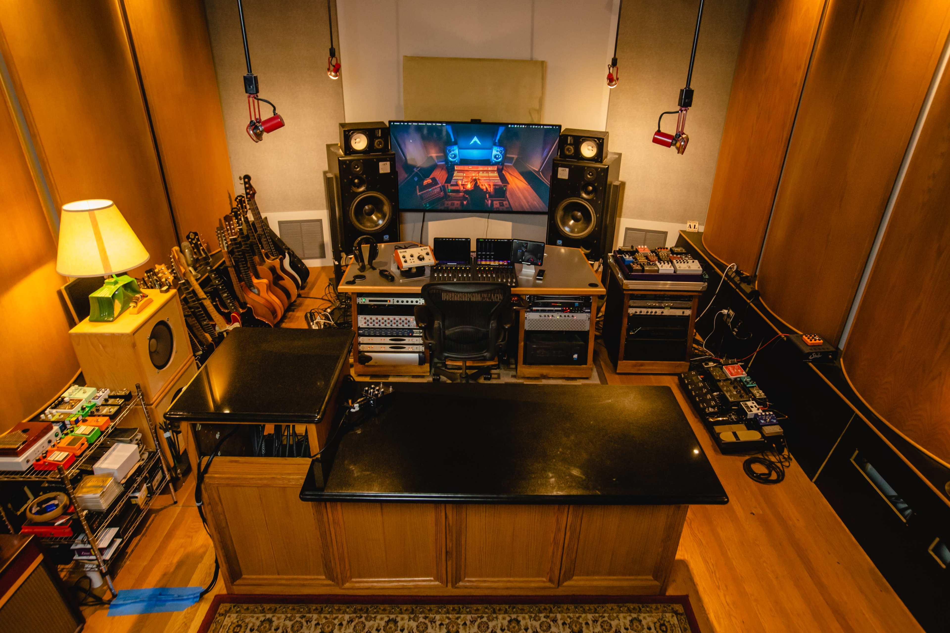 The image shows a recording studio featuring a large central workstation with various equipment and monitors, surrounded by musical instruments and sound gear.