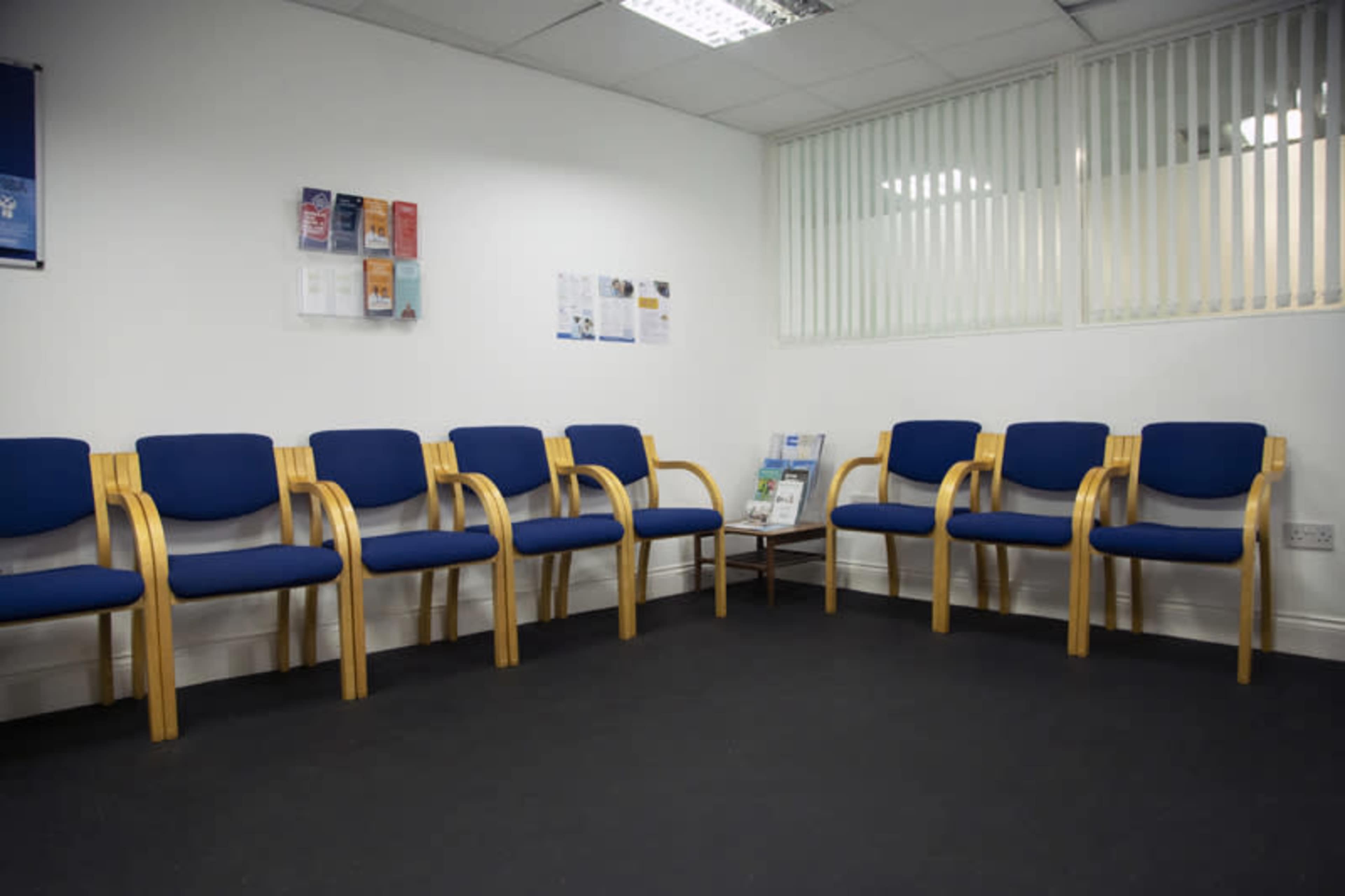 A waiting room features several blue chairs with wooden frames arranged around a small table against a white wall.