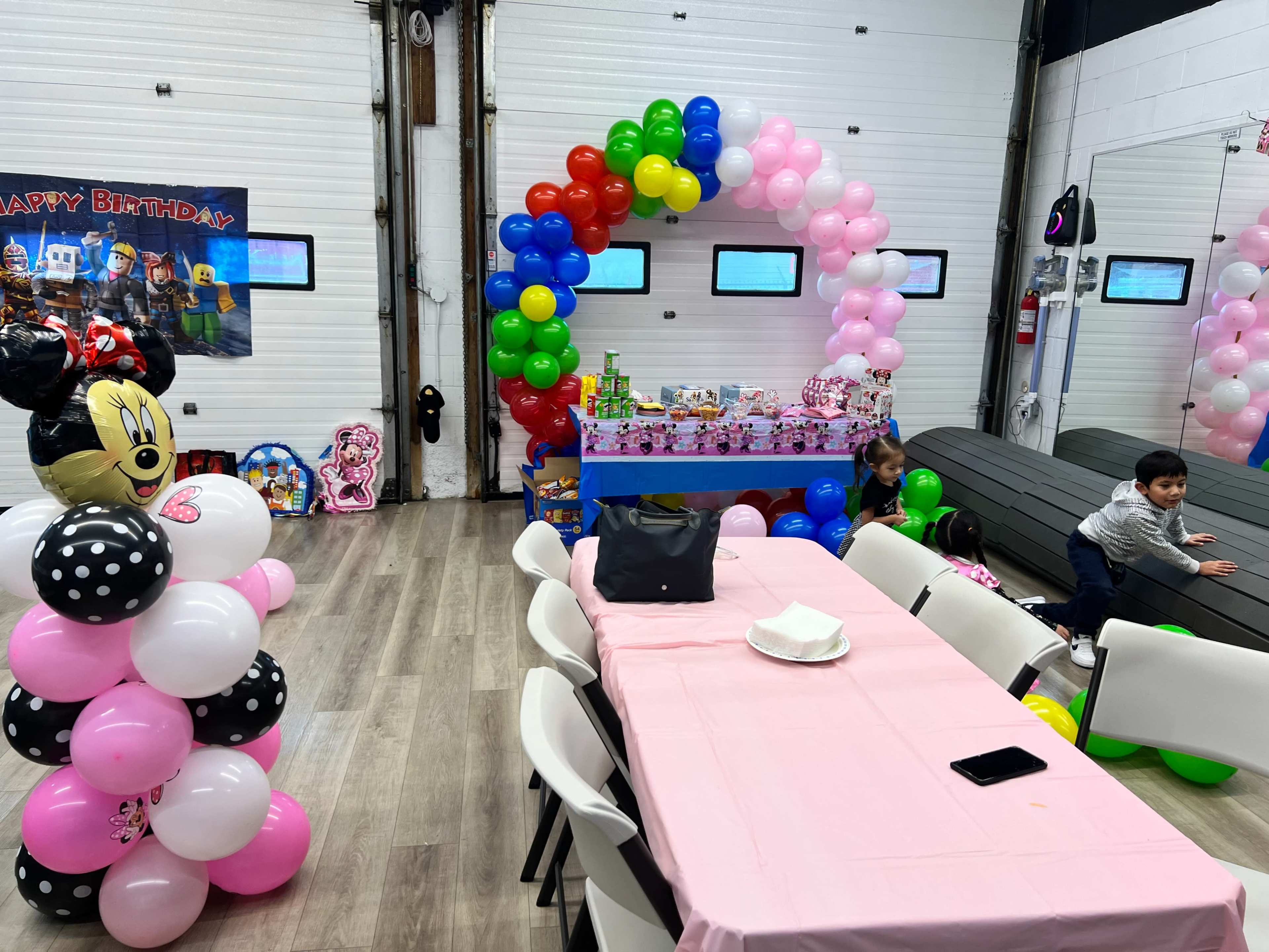 The image shows a decorated party venue featuring colorful balloons, a cake table with gifts, and children playing near the tables.