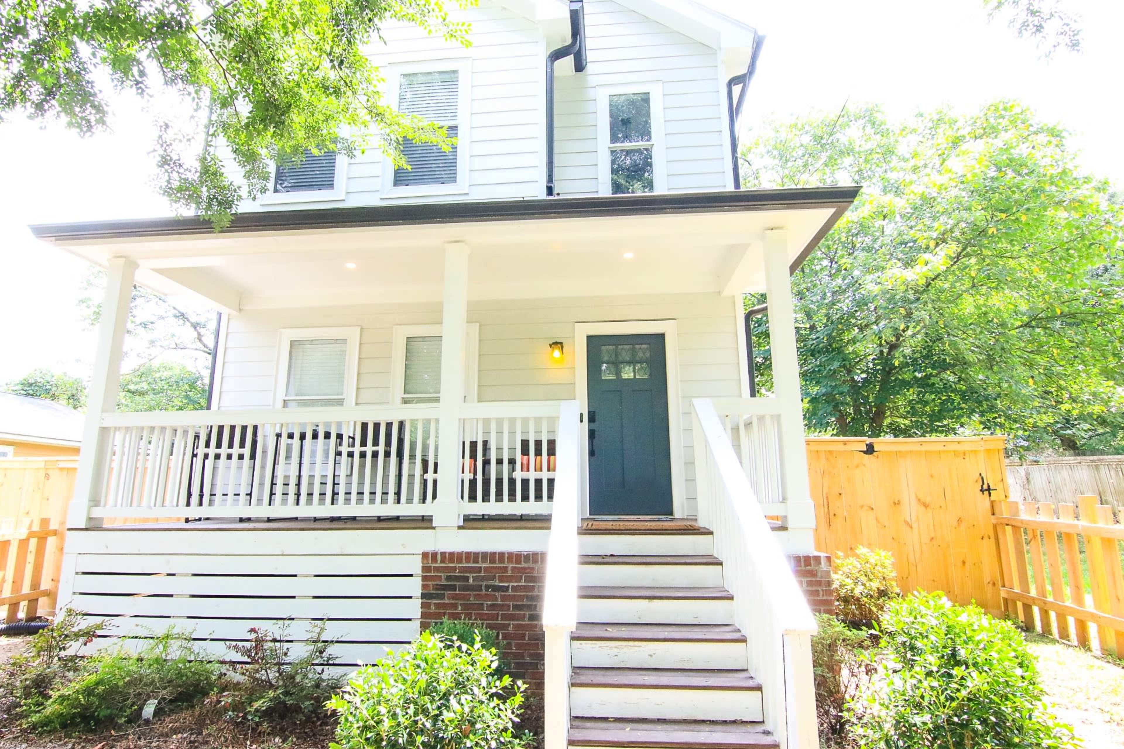 A two-story white house with a blue front door and a porch is surrounded by greenery and a wooden fence.