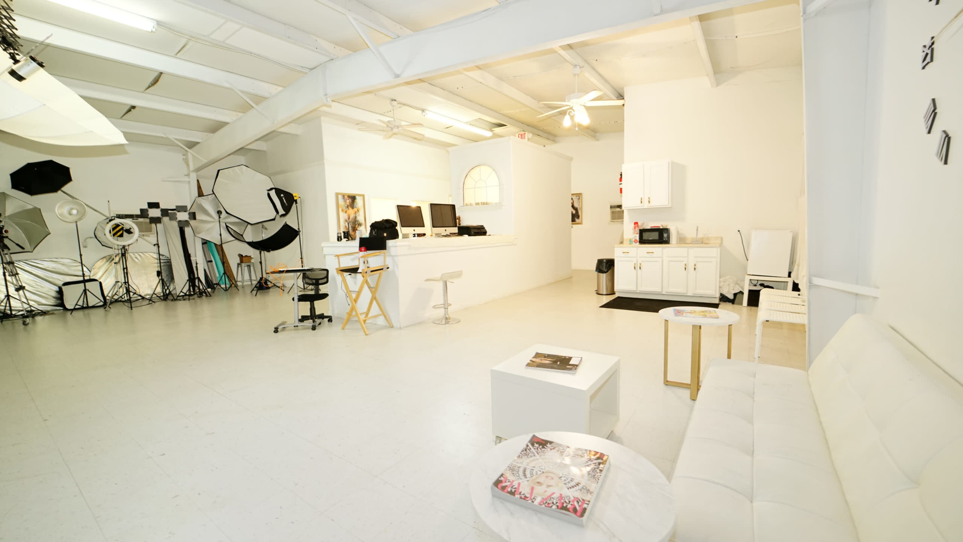 A spacious, well-lit studio featuring a minimalist seating area, a kitchenette, and various photography equipment in the background.