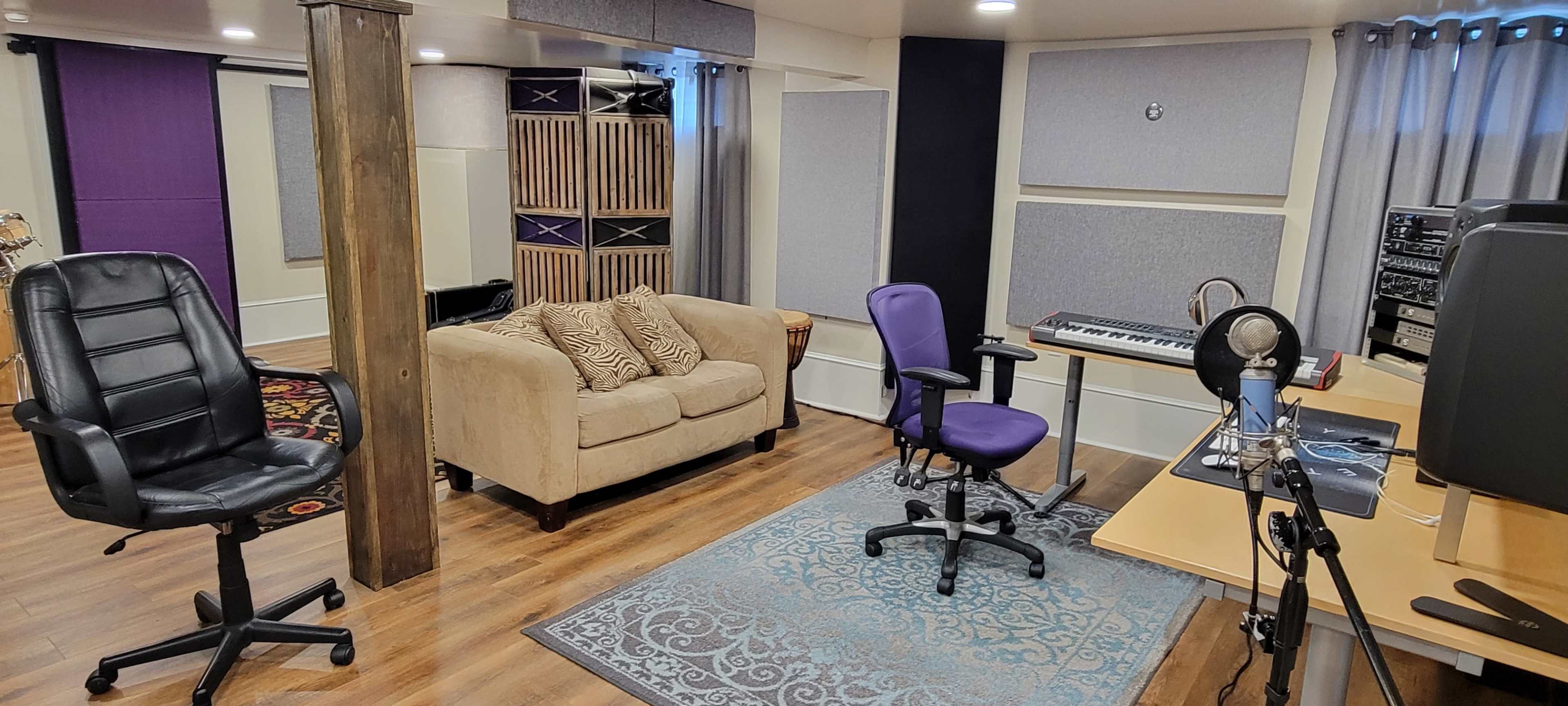 A music recording studio with a beige couch, two office chairs, a keyboard on a desk, and soundproof panels on the walls.