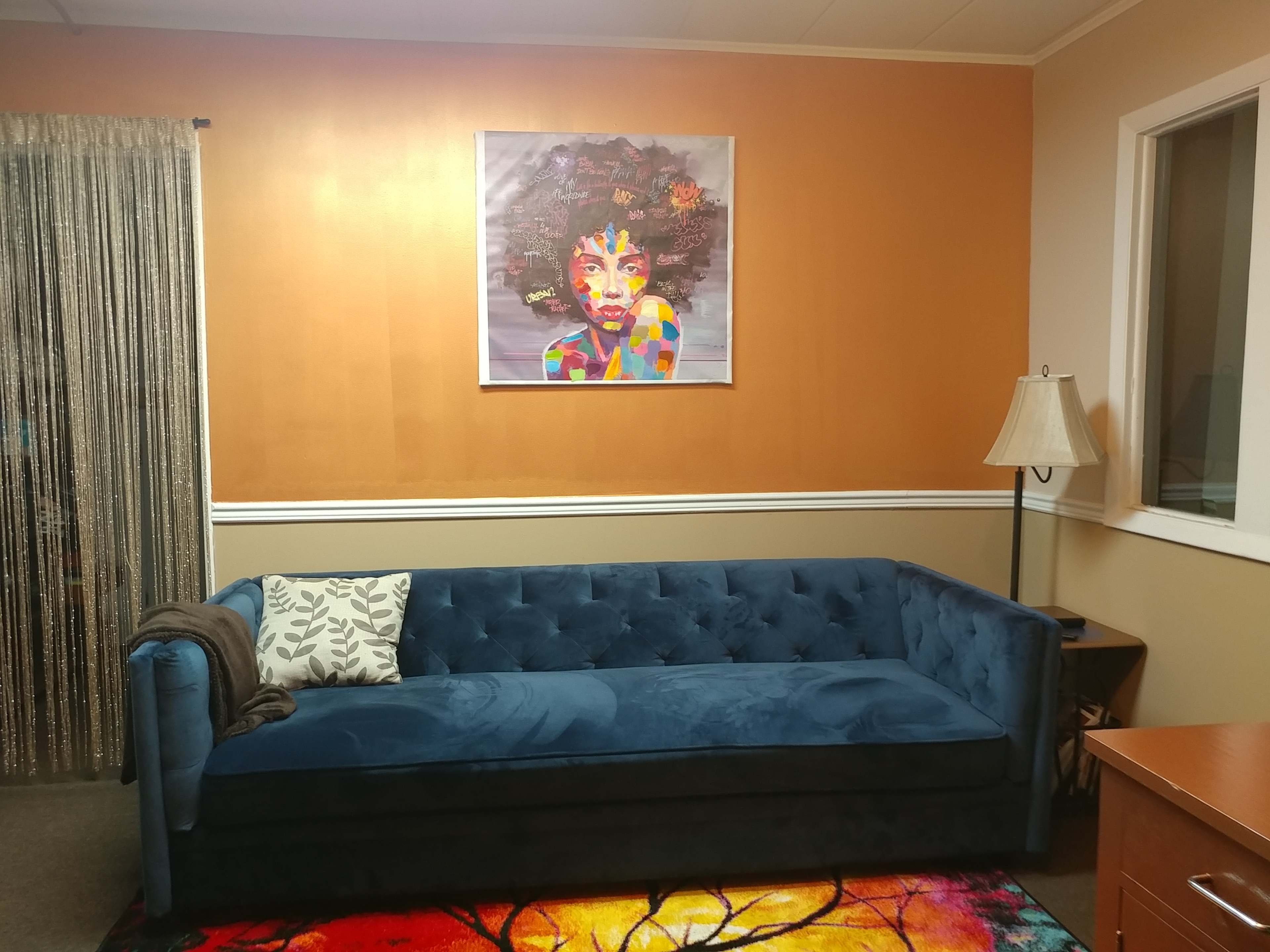 The image shows a cozy room featuring a blue velvet sofa, a colorful artwork on the wall, and a lamp beside a wooden side table.