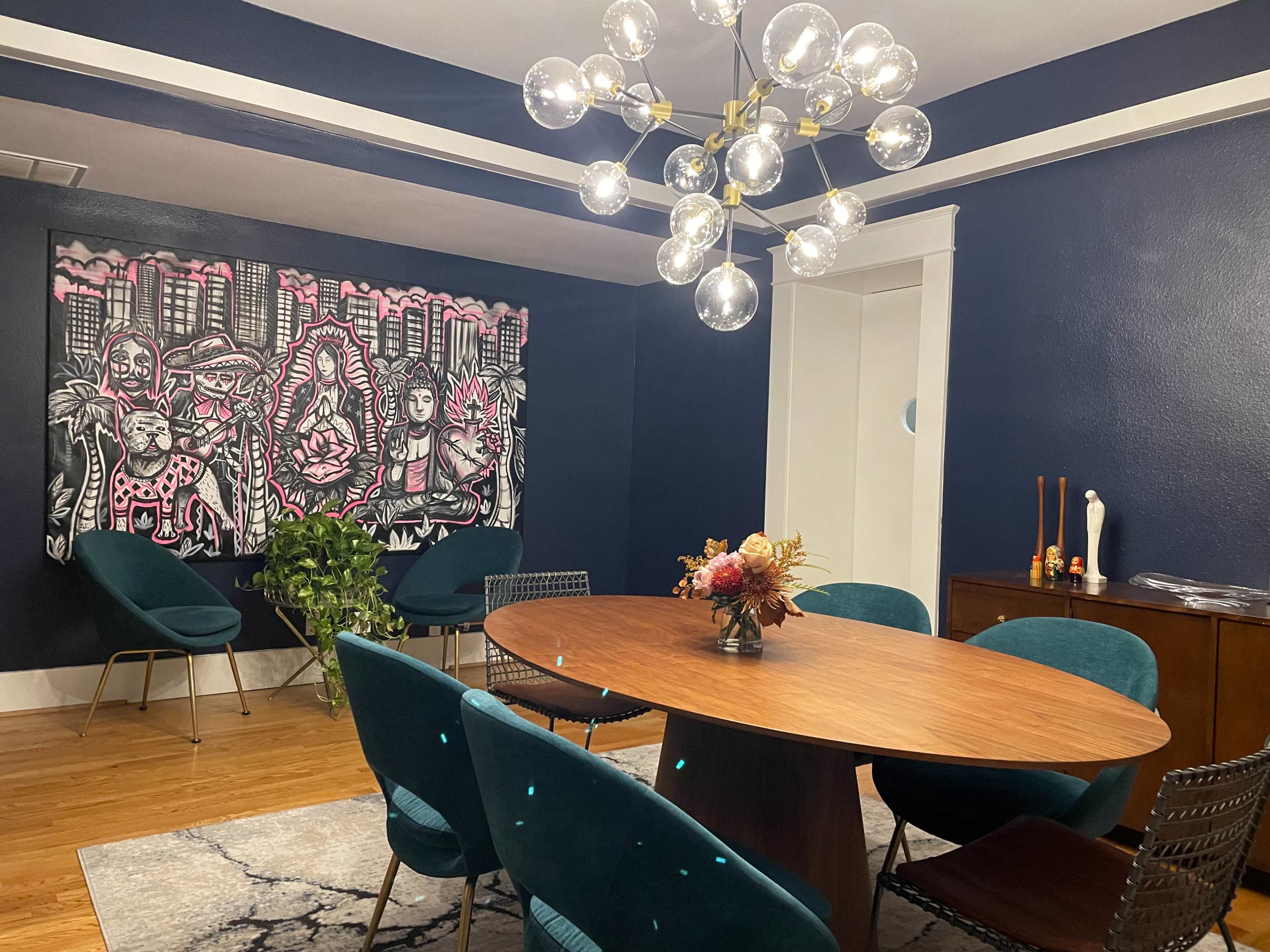 A dining room features a round wooden table surrounded by teal chairs, with a large mural on the wall and a modern chandelier overhead.