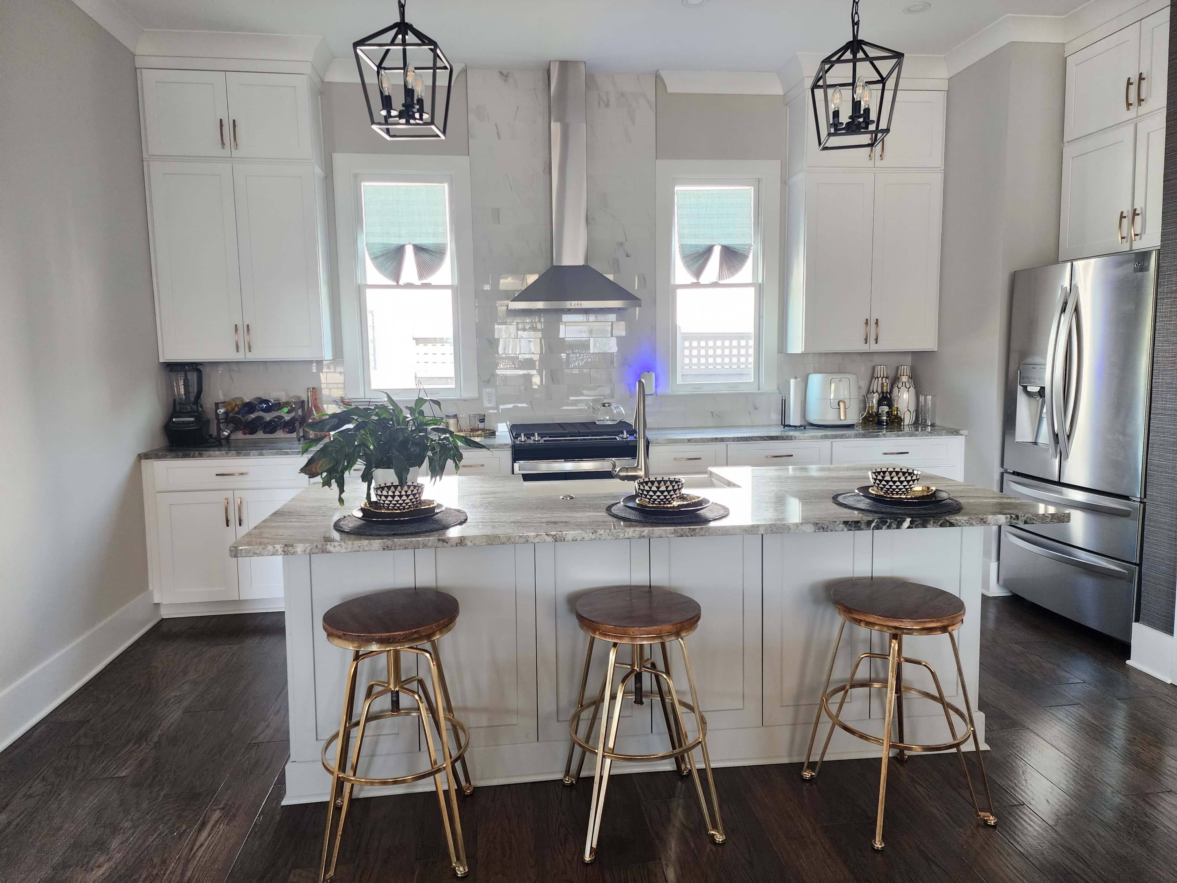 A modern kitchen features a central island with three metal bar stools, pendant lighting, and stainless steel appliances.