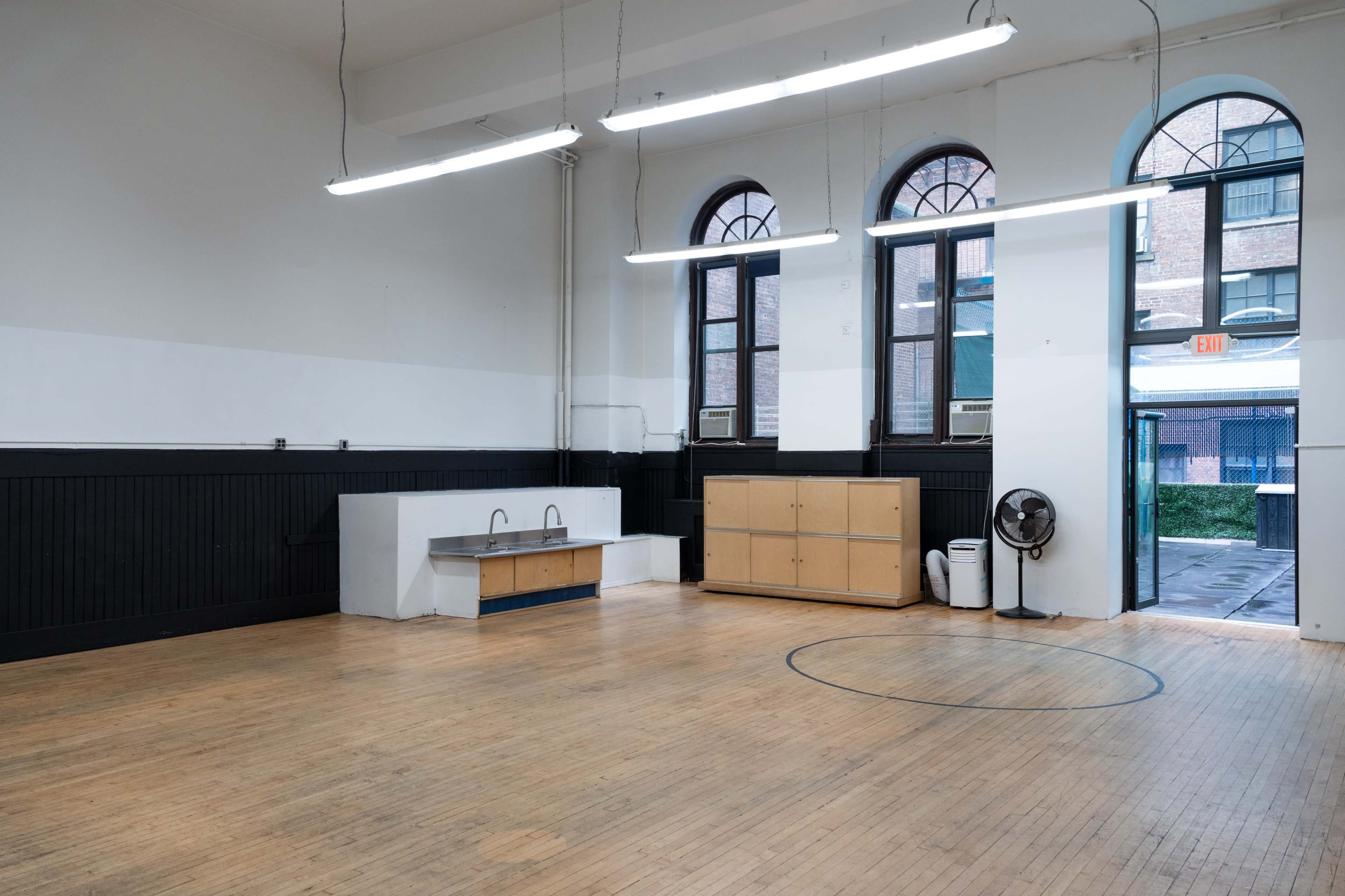 The image shows a large, empty room with hardwood flooring, a white sink against one wall, a wooden cabinet, and large windows that allow natural light to enter.