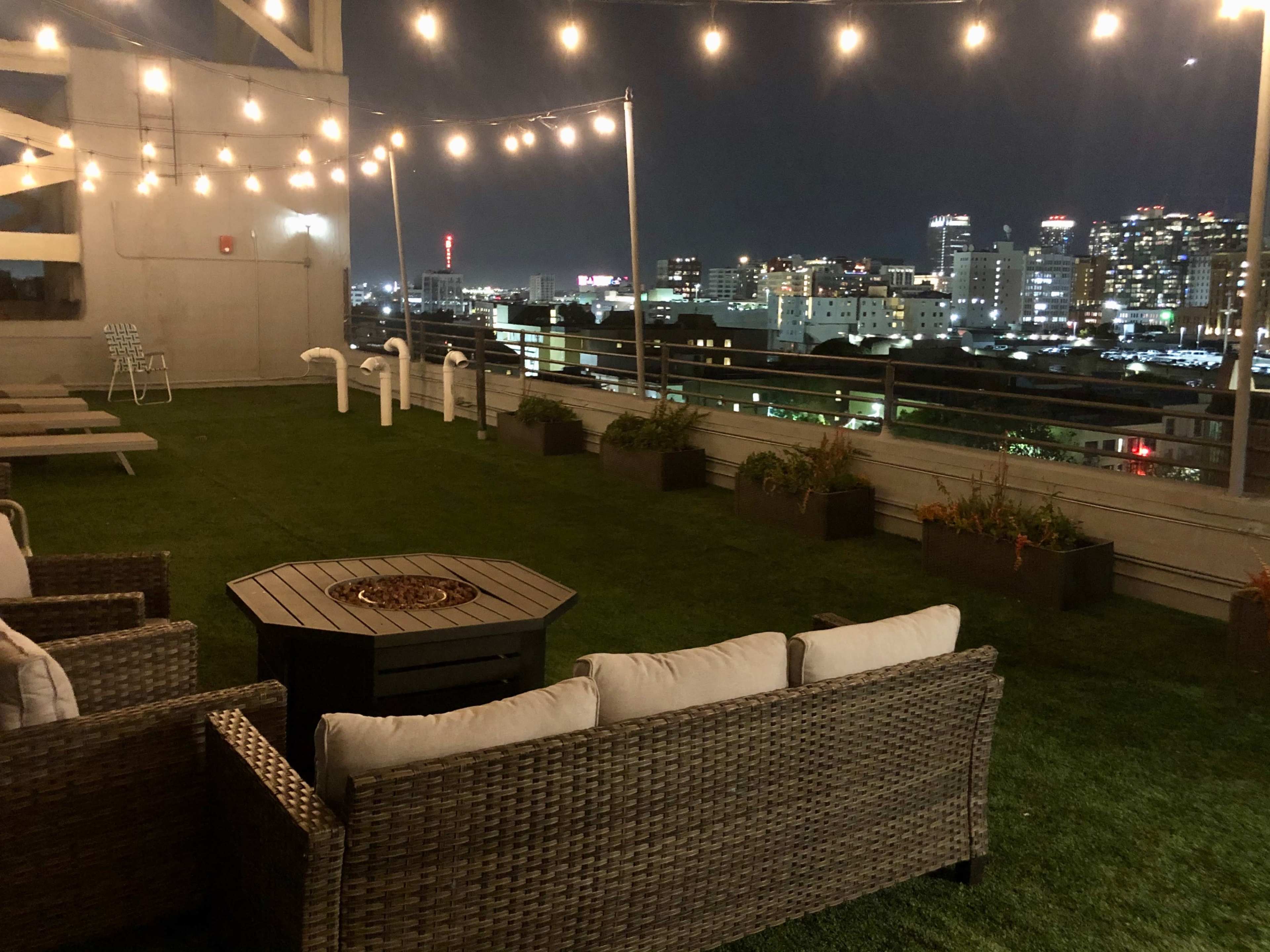 A rooftop terrace with outdoor seating, a fire pit, and string lights, overlooking a city skyline at night.