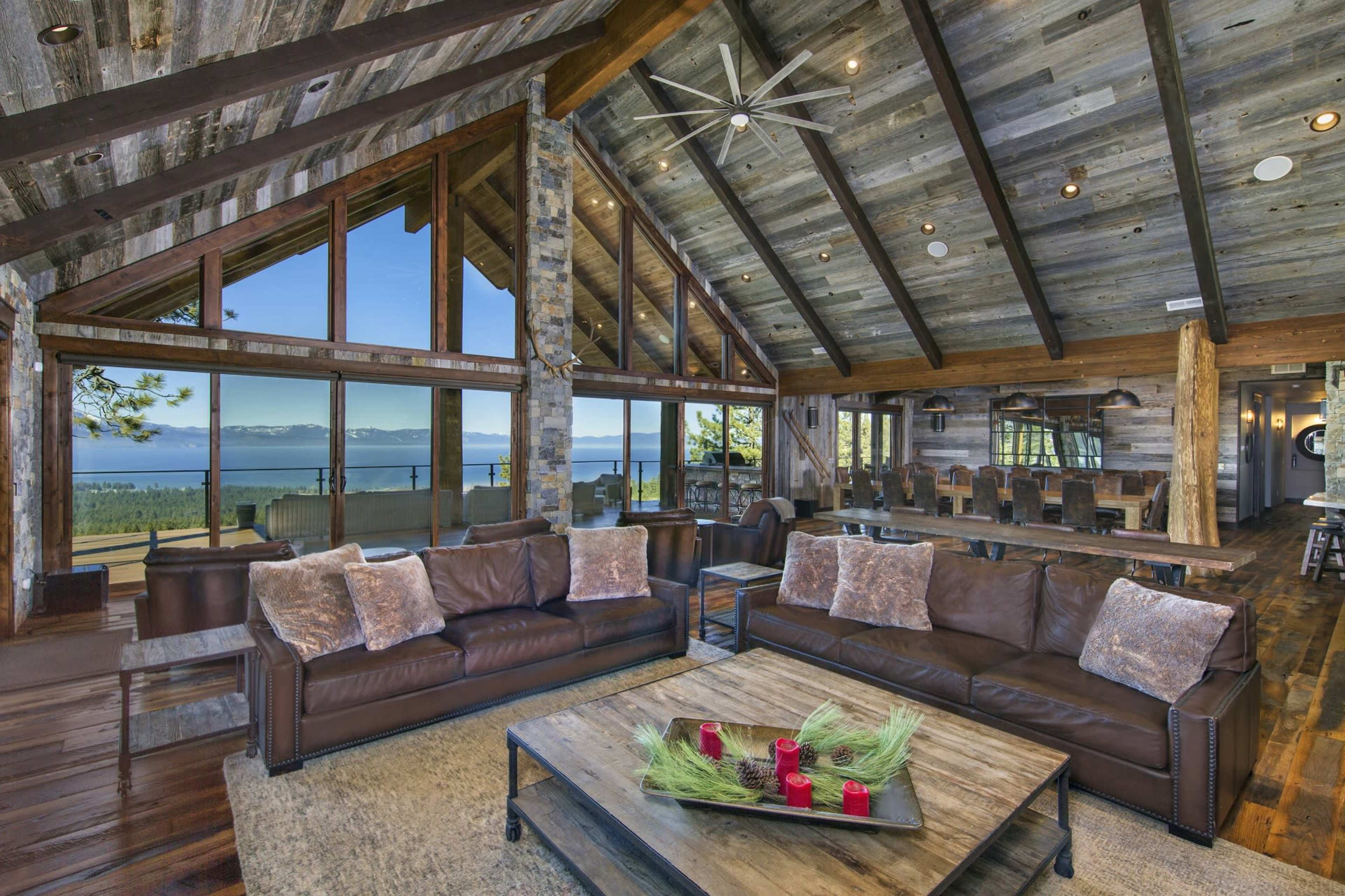 A spacious living area features leather sofas, a wooden table with decorative elements, and large windows showcasing a view of the lake and mountains.