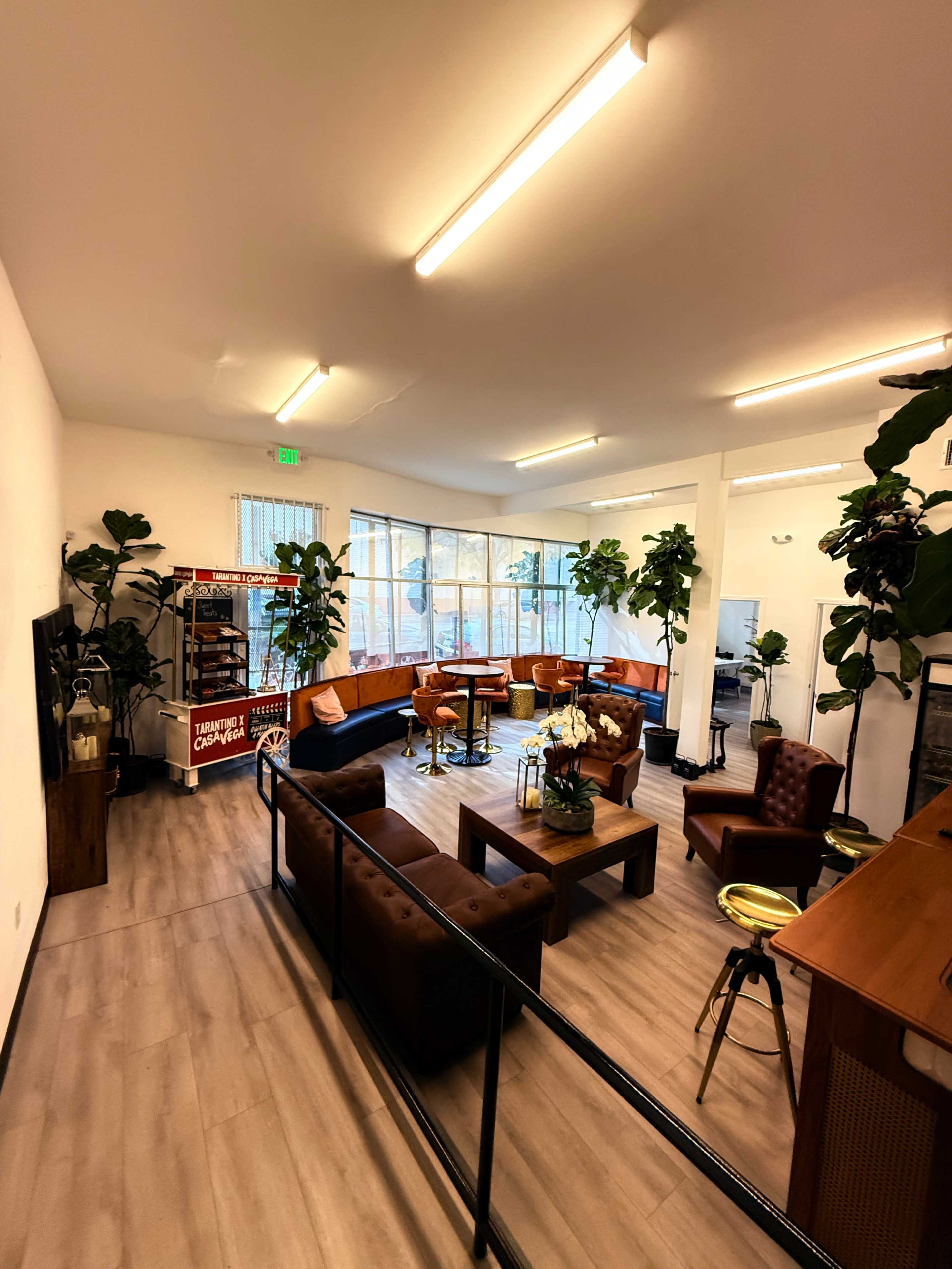 The image shows a spacious lounge area with comfortable seating, a wooden table, and large windows that allow natural light to enter, surrounded by indoor plants.