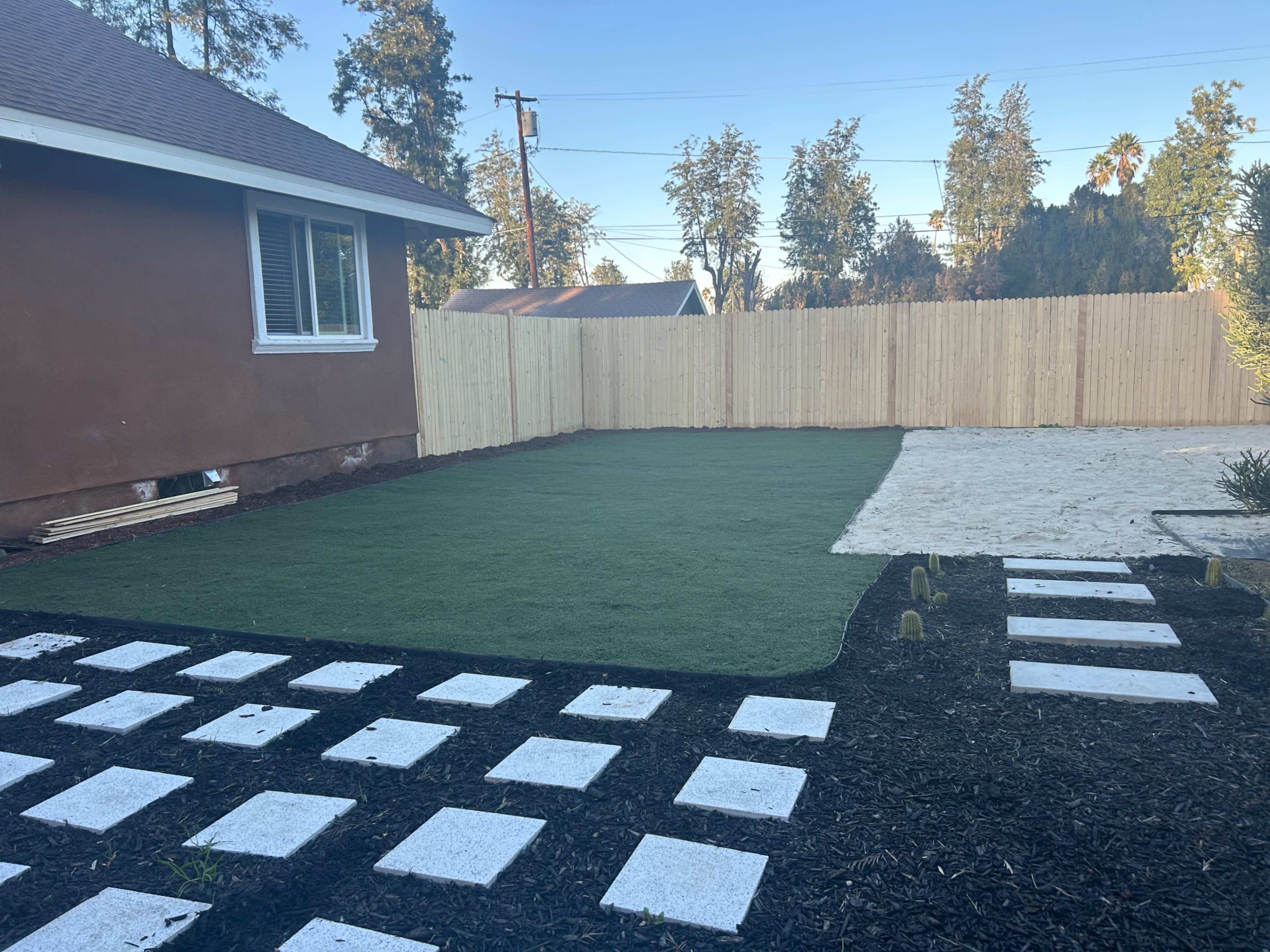 A yard with artificial grass on one side and a sandy area on the other, bordered by a wooden fence and pavement made of white square stones.