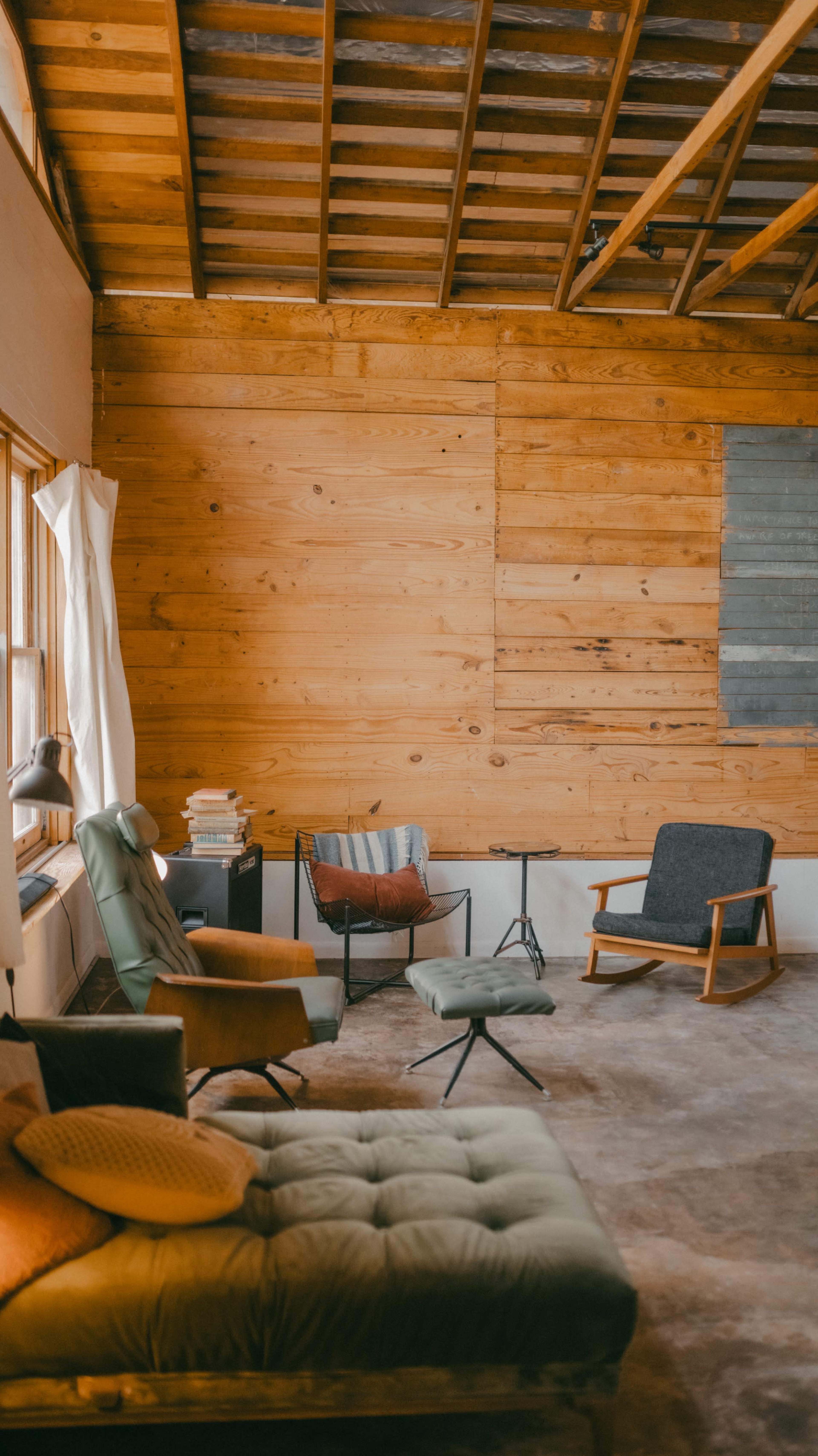 The image shows a minimalist living space with wooden walls and a mix of modern and vintage furniture, including a green armchair, a black chair, and a cushioned sofa.