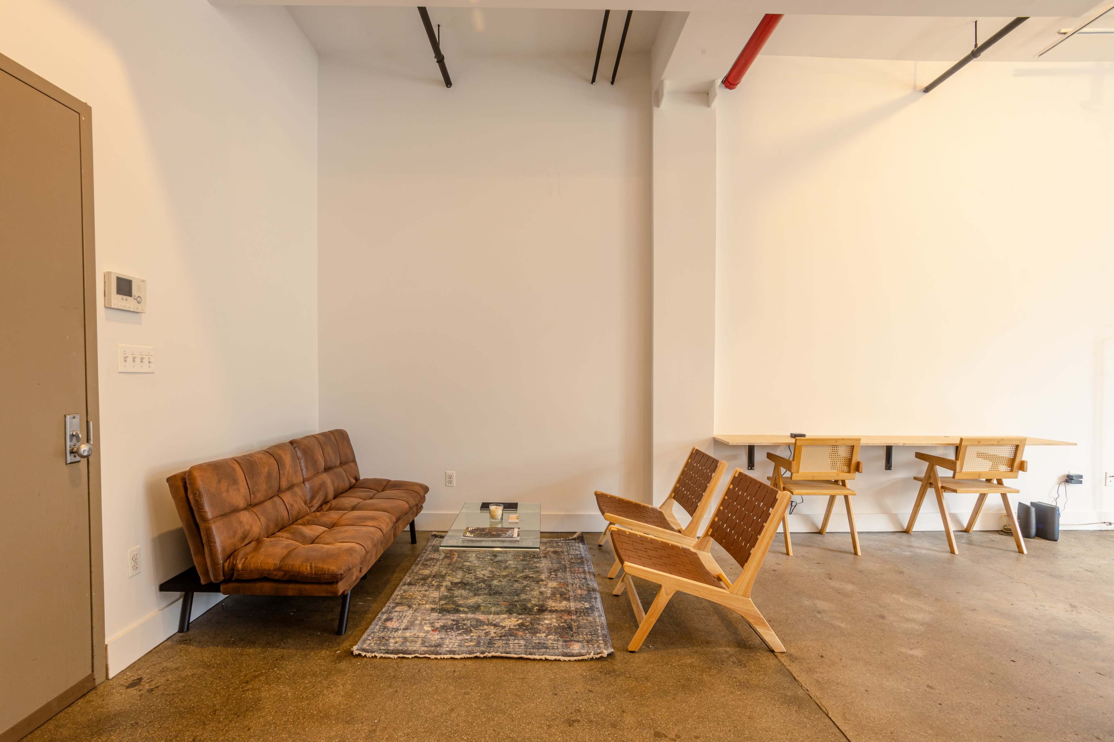 A minimalist interior featuring a brown leather sofa, two wooden chairs, a rug, and a table against a plain white wall.
