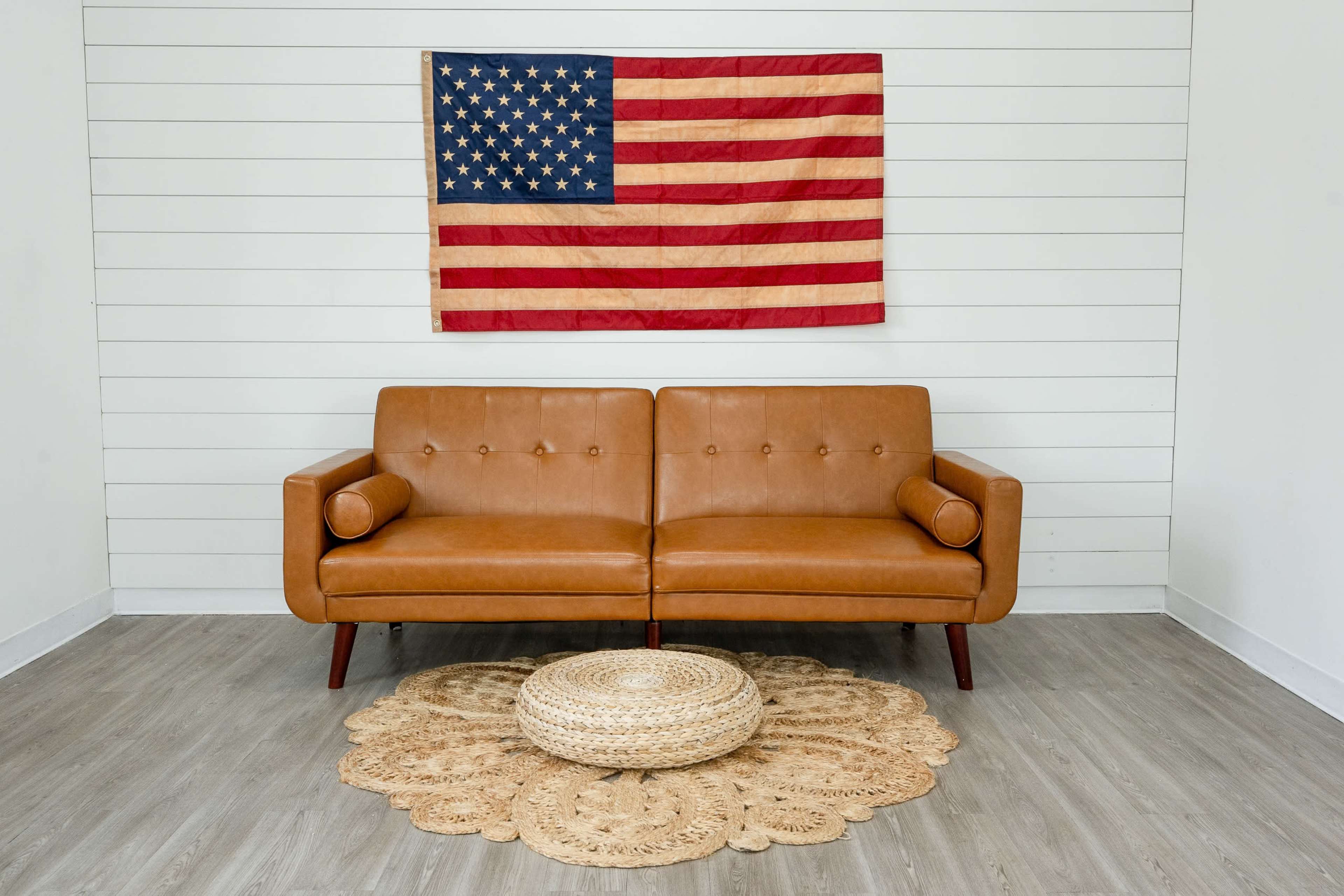 A brown leather sofa is positioned in front of a wooden wall, adorned with a large American flag, and a round woven rug is placed on the floor underneath a coffee table.