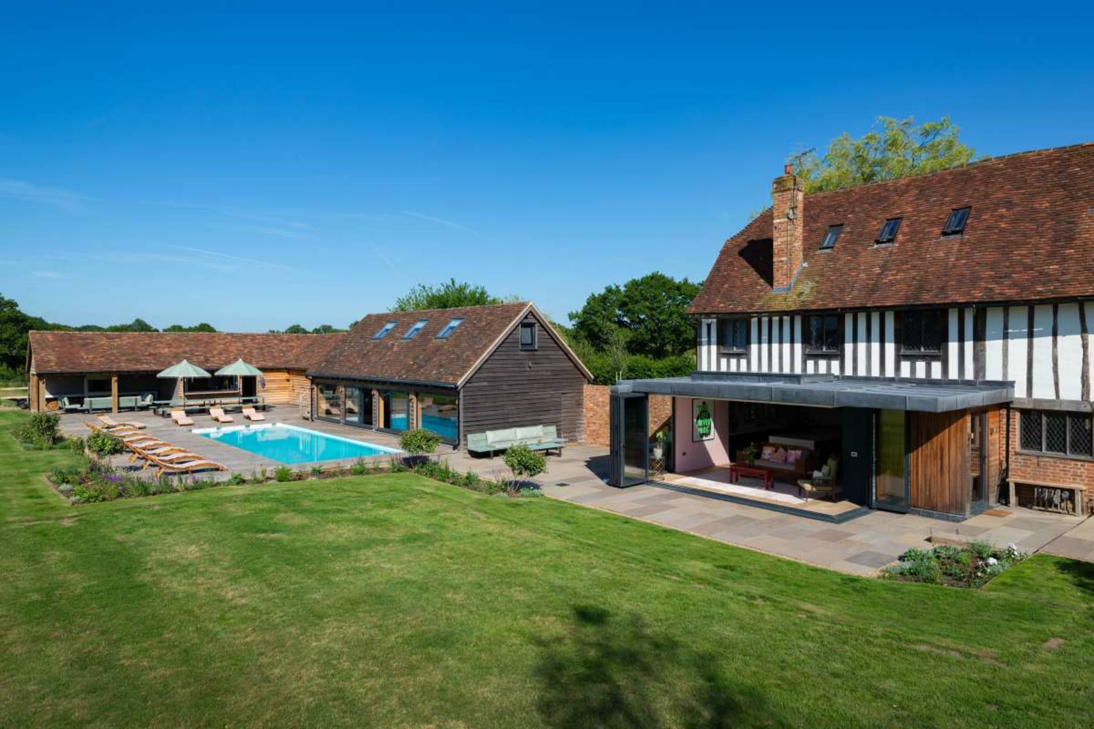 The image shows a modern house with a large pool surrounded by lounge chairs, set in a green landscape with blue skies.