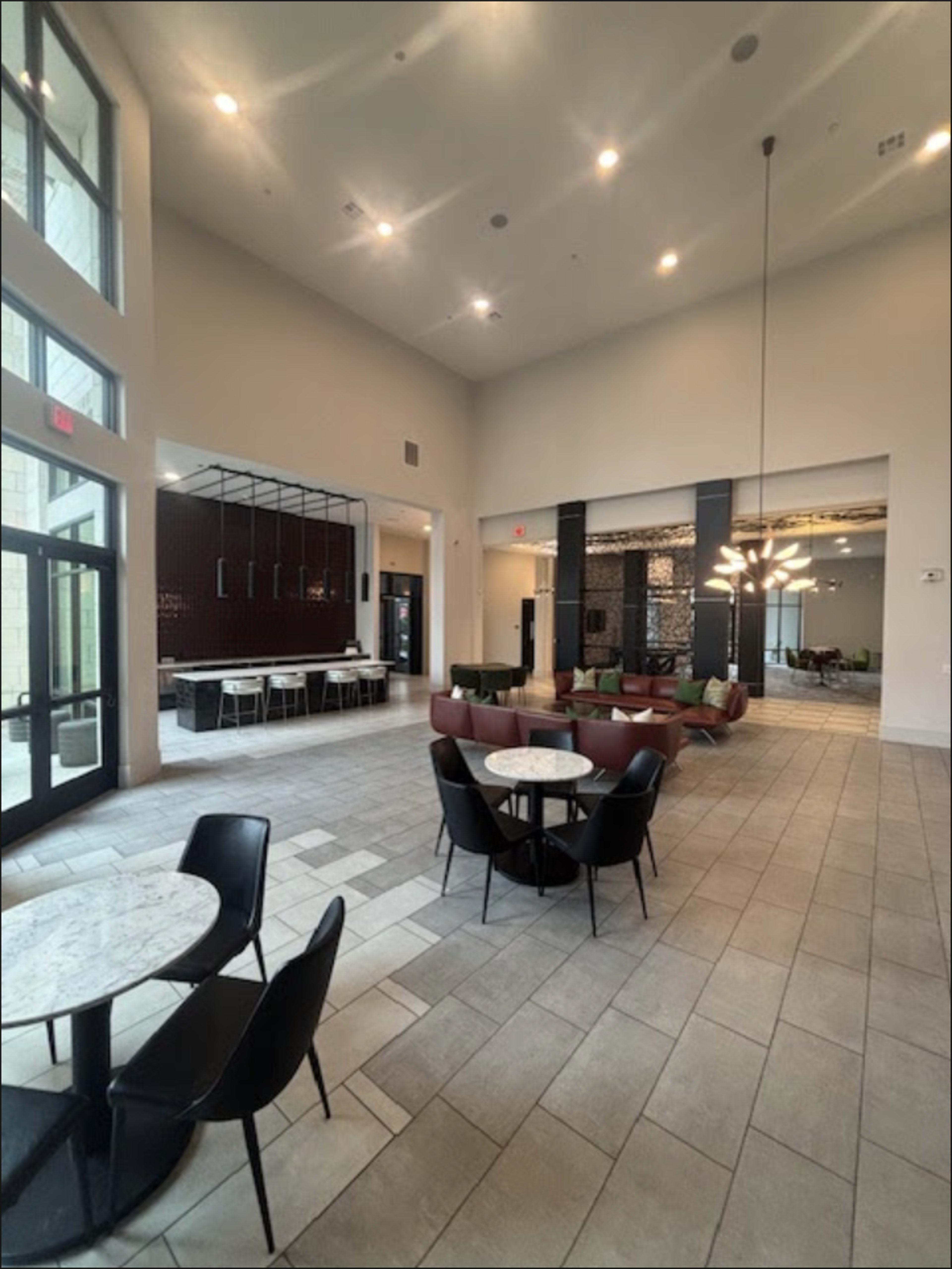 The image shows a spacious, modern interior with high ceilings, featuring a seating area with marble tables and black chairs, alongside a dining area with a long table and contemporary lighting fixtures.