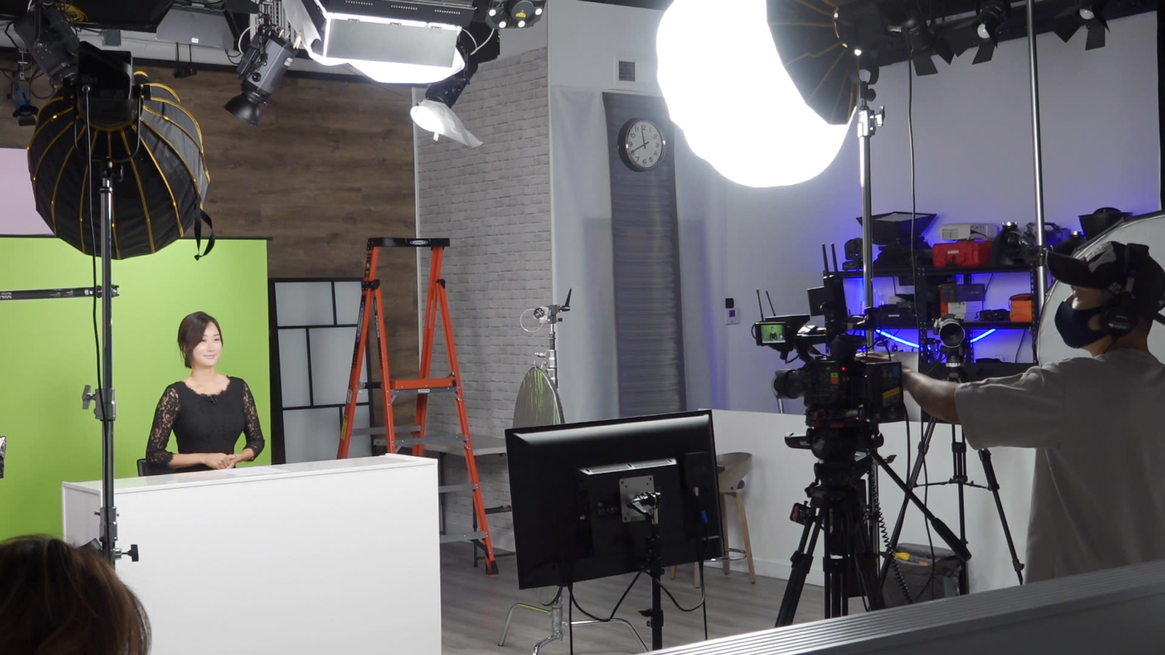 A woman stands behind a white desk in a studio set up for a television broadcast, with cameras and lighting equipment arranged around her.