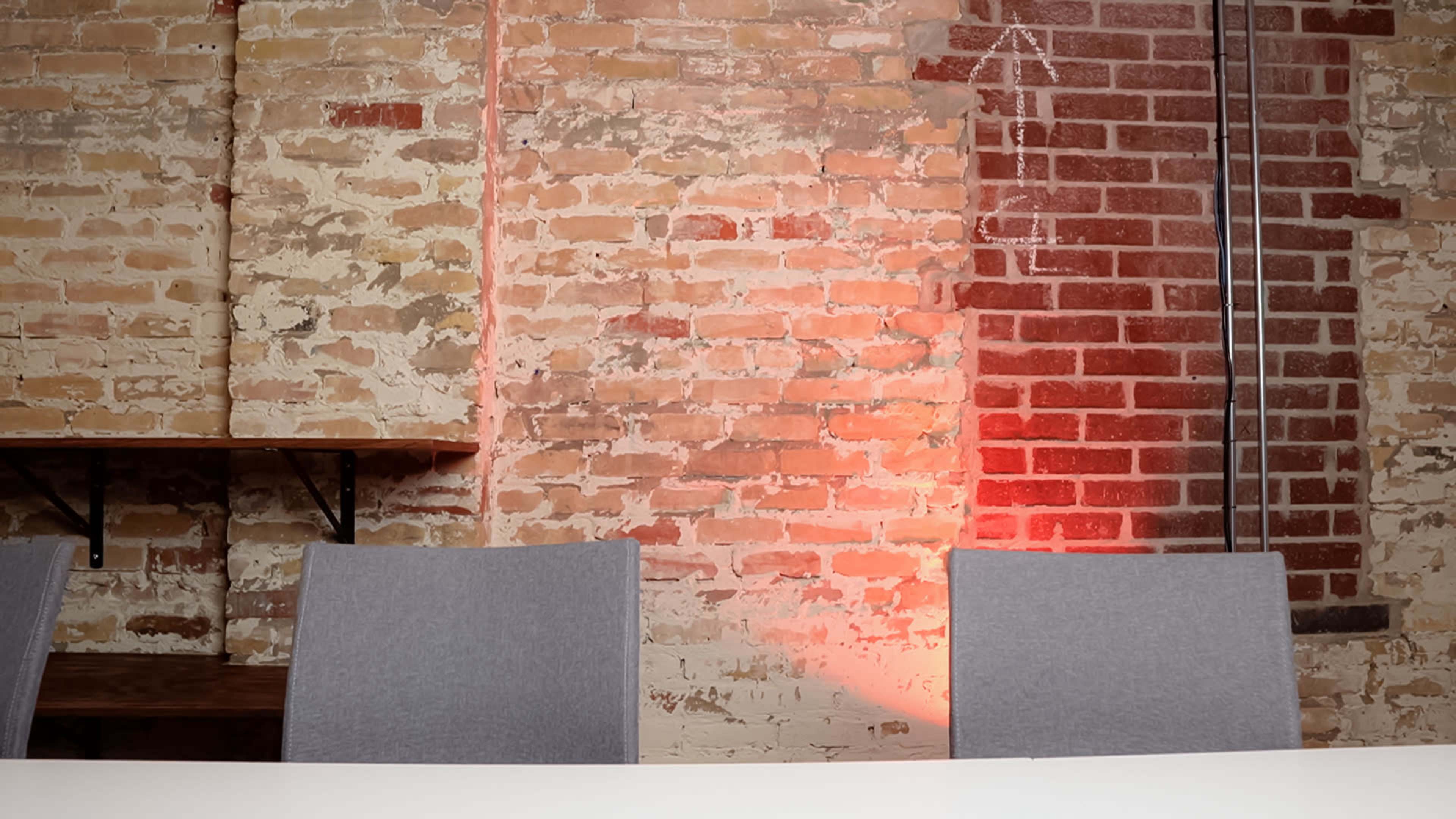 A table with empty gray chairs set against a textured brick wall, partially illuminated by a red light.