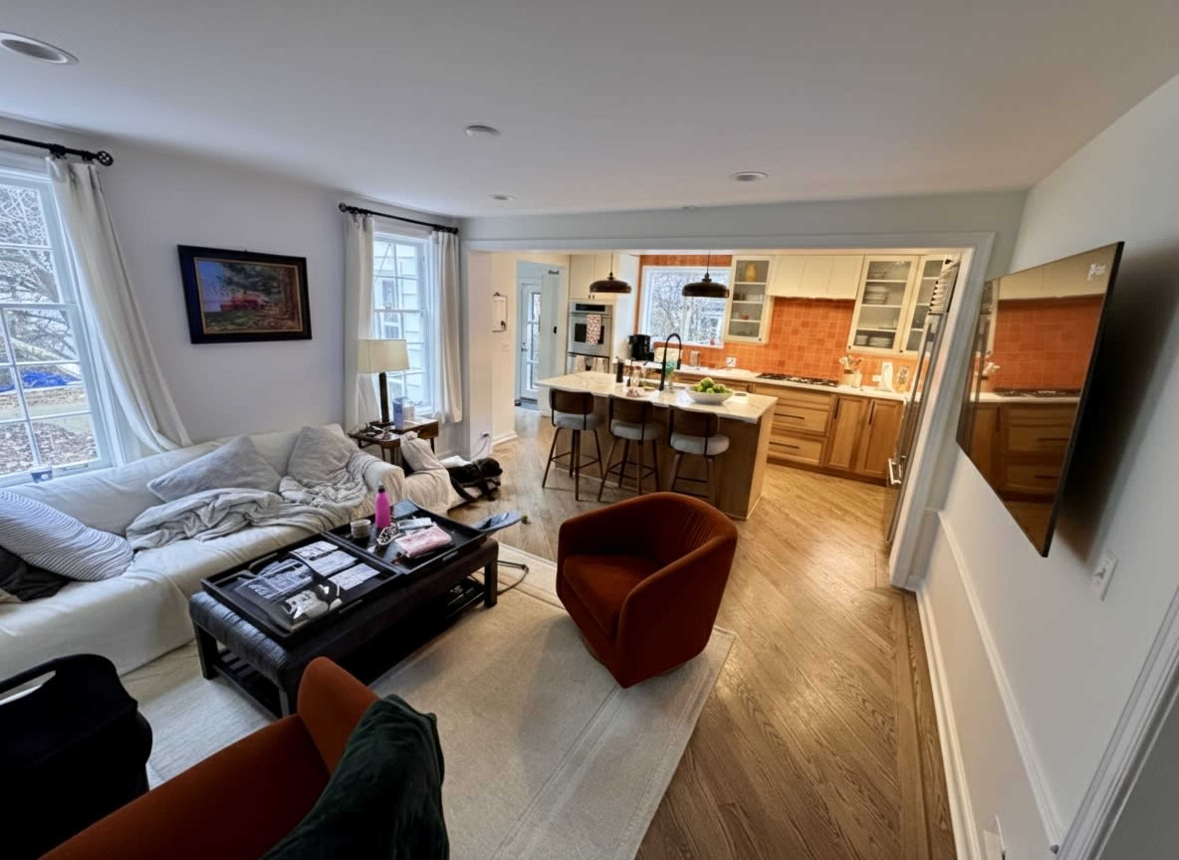 The image shows a modern living room with a white sofa, coffee table, and a view of a kitchen area featuring orange tiled walls and wooden cabinets.