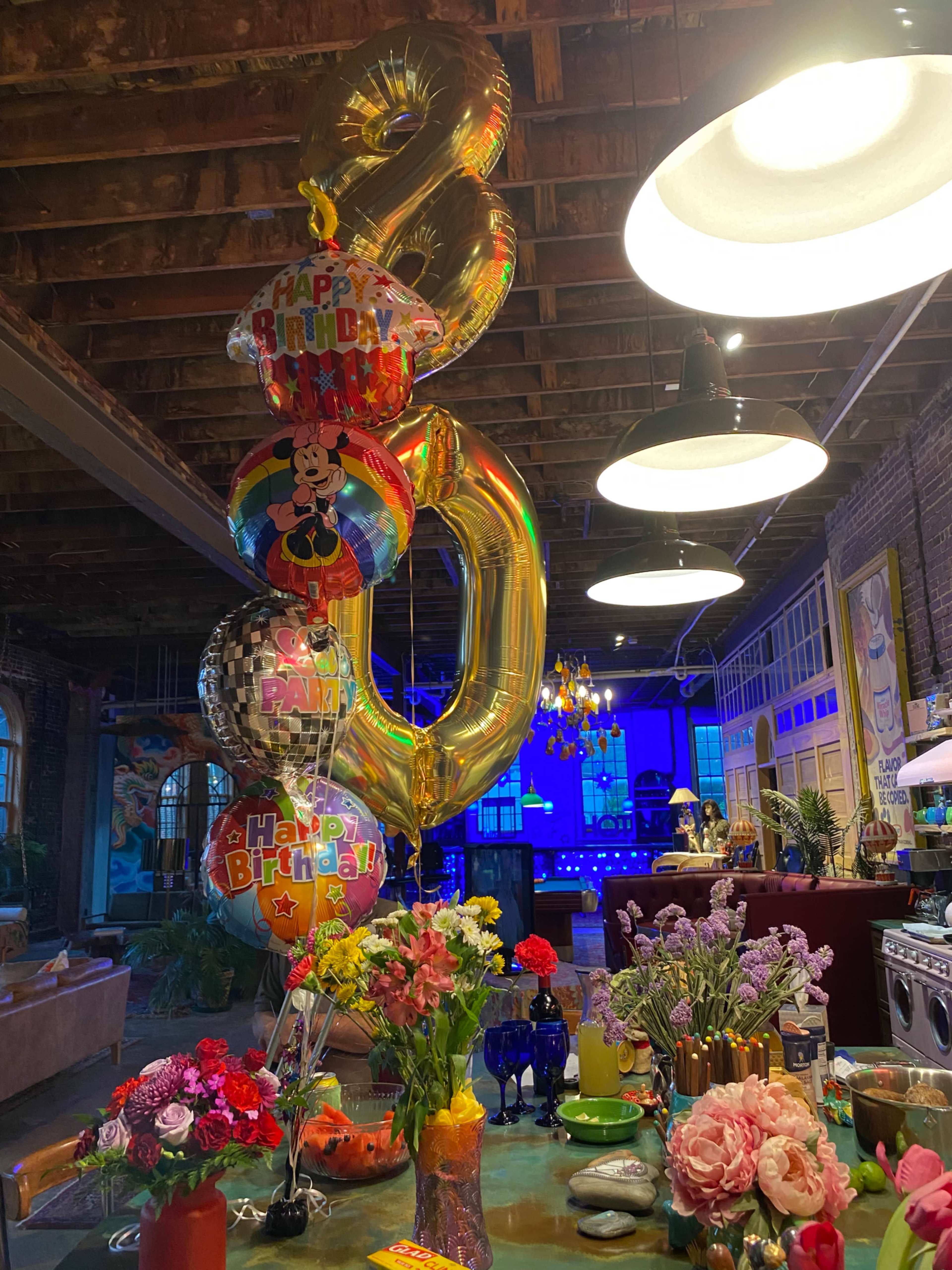 The image shows a decorated interior with a large "8" balloon, colorful birthday balloons, and a table adorned with various flowers and vibrant tableware.