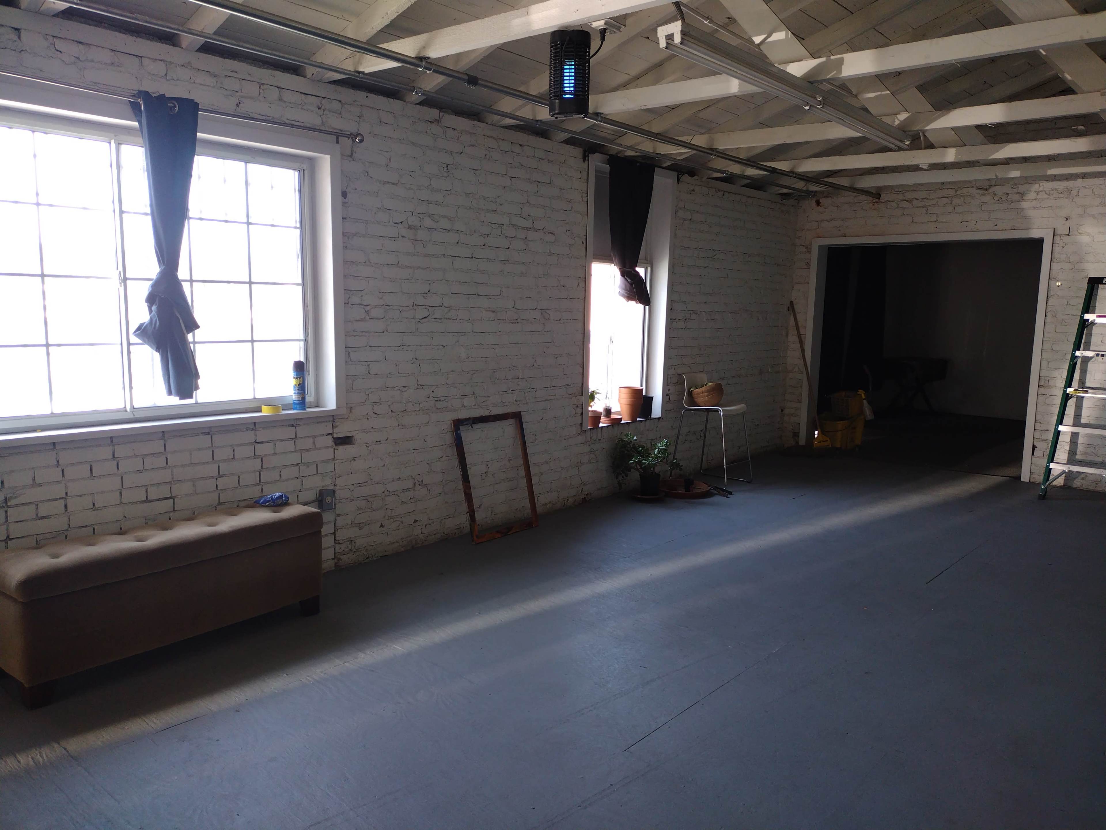 A spacious room with white brick walls, two large windows with curtains, a bench, and various plants along with a ladder and other furnishings in the background.