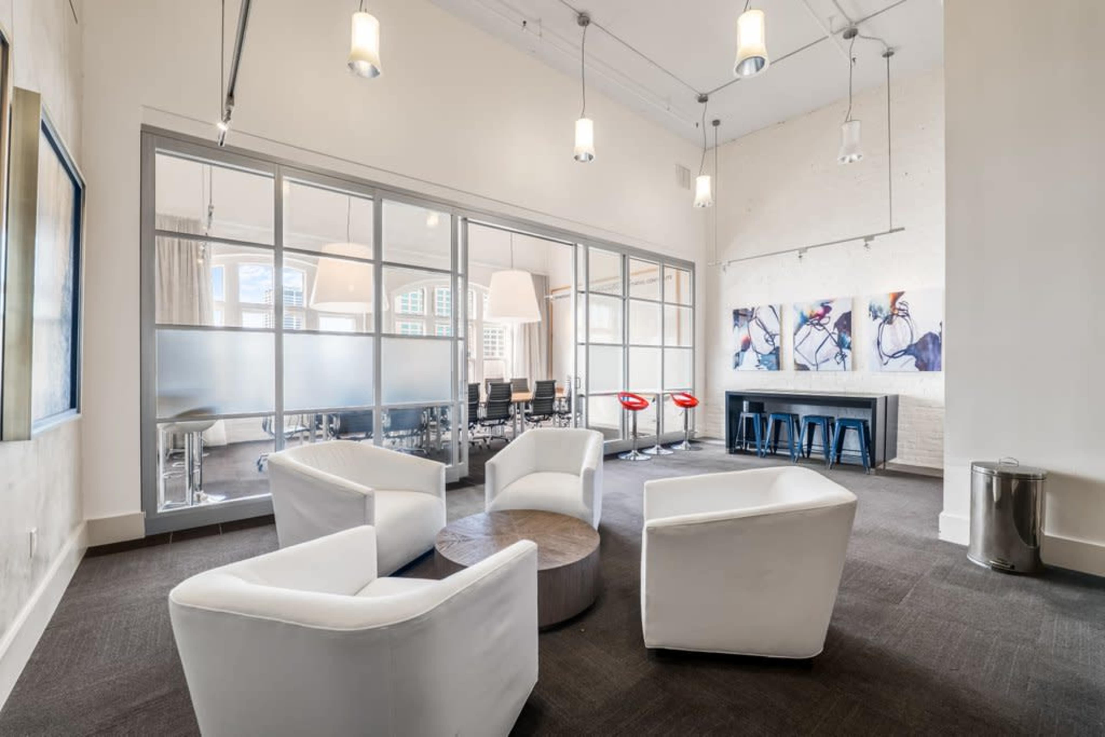 The image shows a modern office interior with a seating area featuring white armchairs surrounding a circular wooden table, adjacent to a glass-walled conference room and artwork on the wall.