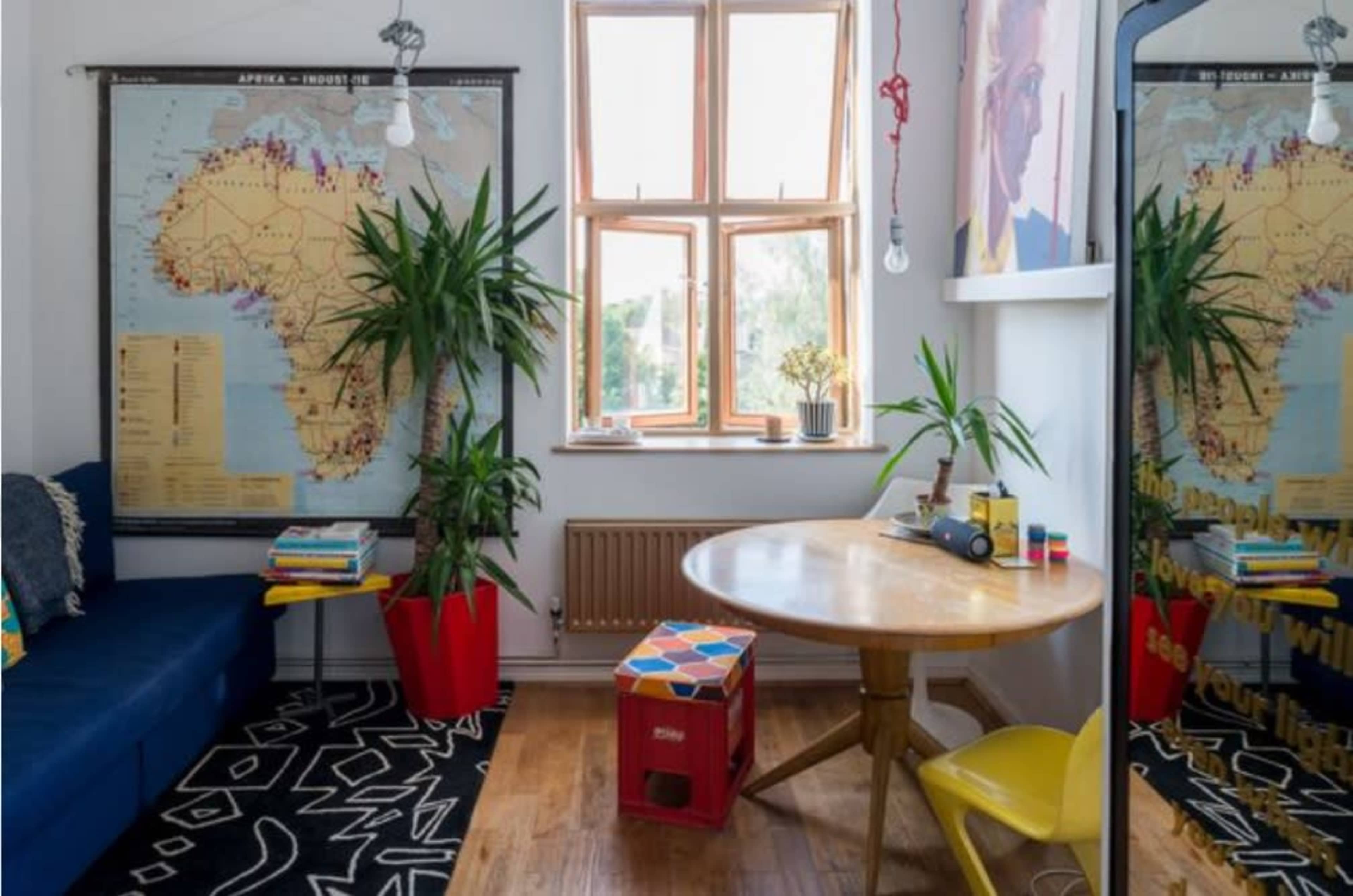 The image shows a bright room featuring a map of Africa on the wall, a round wooden table with colorful items, and a cozy seating area with plants and books.
