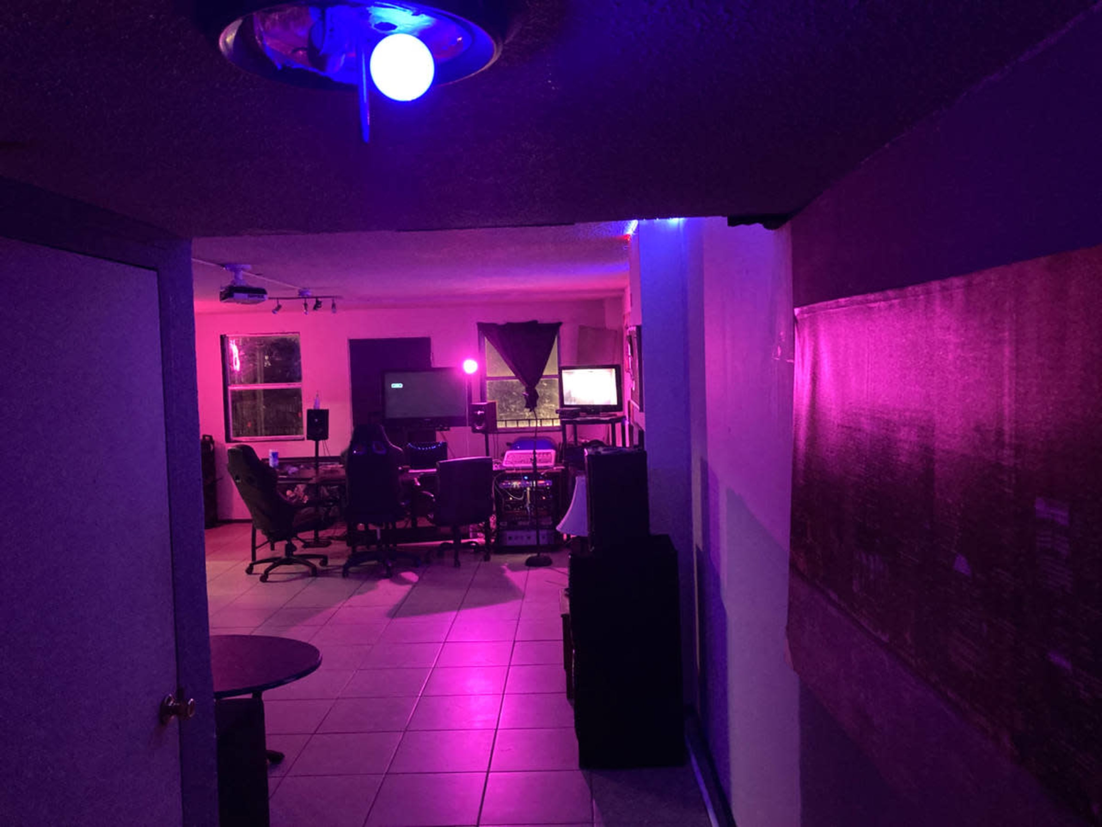 A dimly lit hallway leads to a room filled with purple ambient lighting, featuring a seating area and screens.