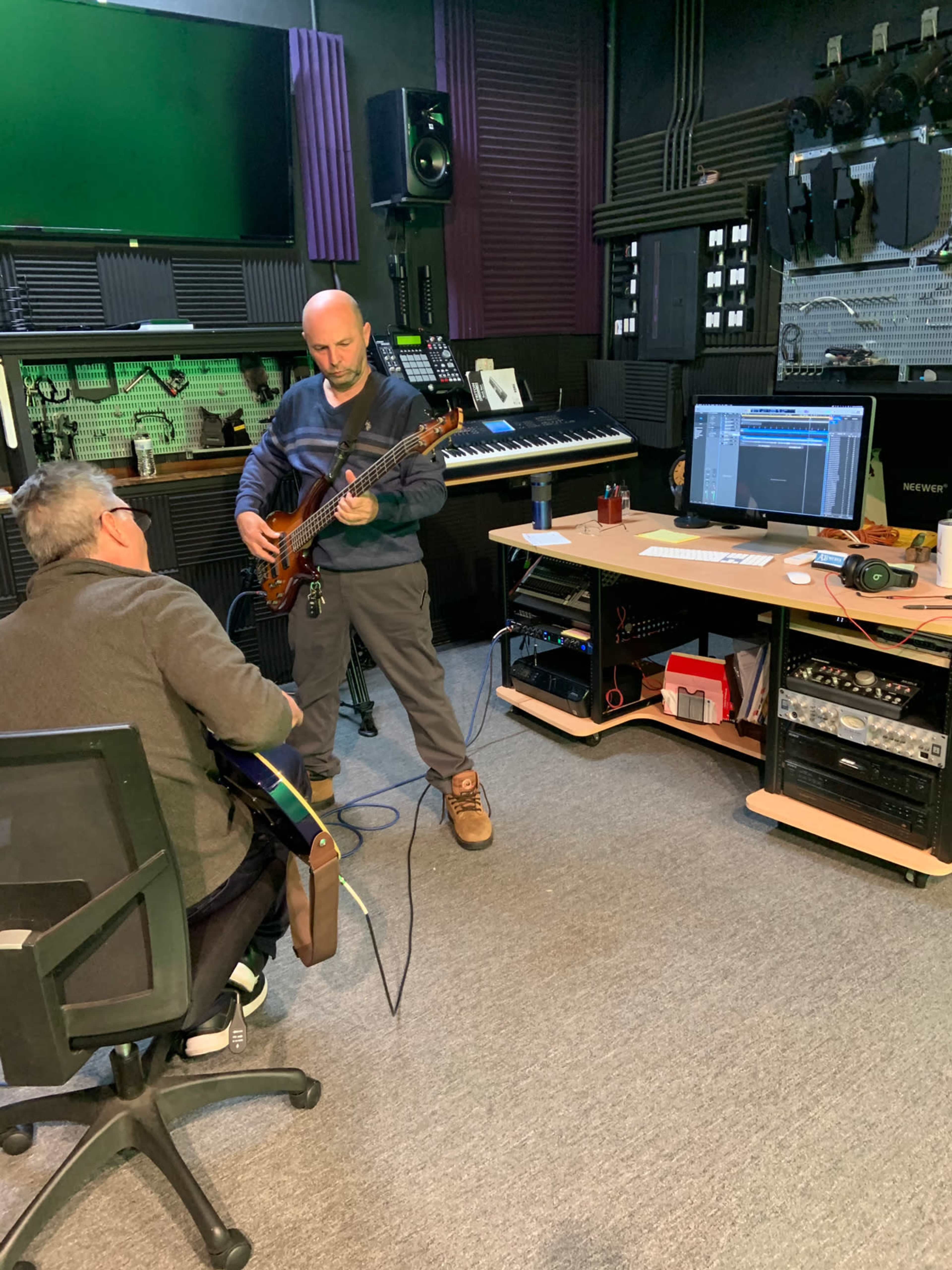 Two musicians are collaborating in a recording studio, with one playing an electric bass and the other seated with an acoustic guitar, while a computer screen displays music production software.