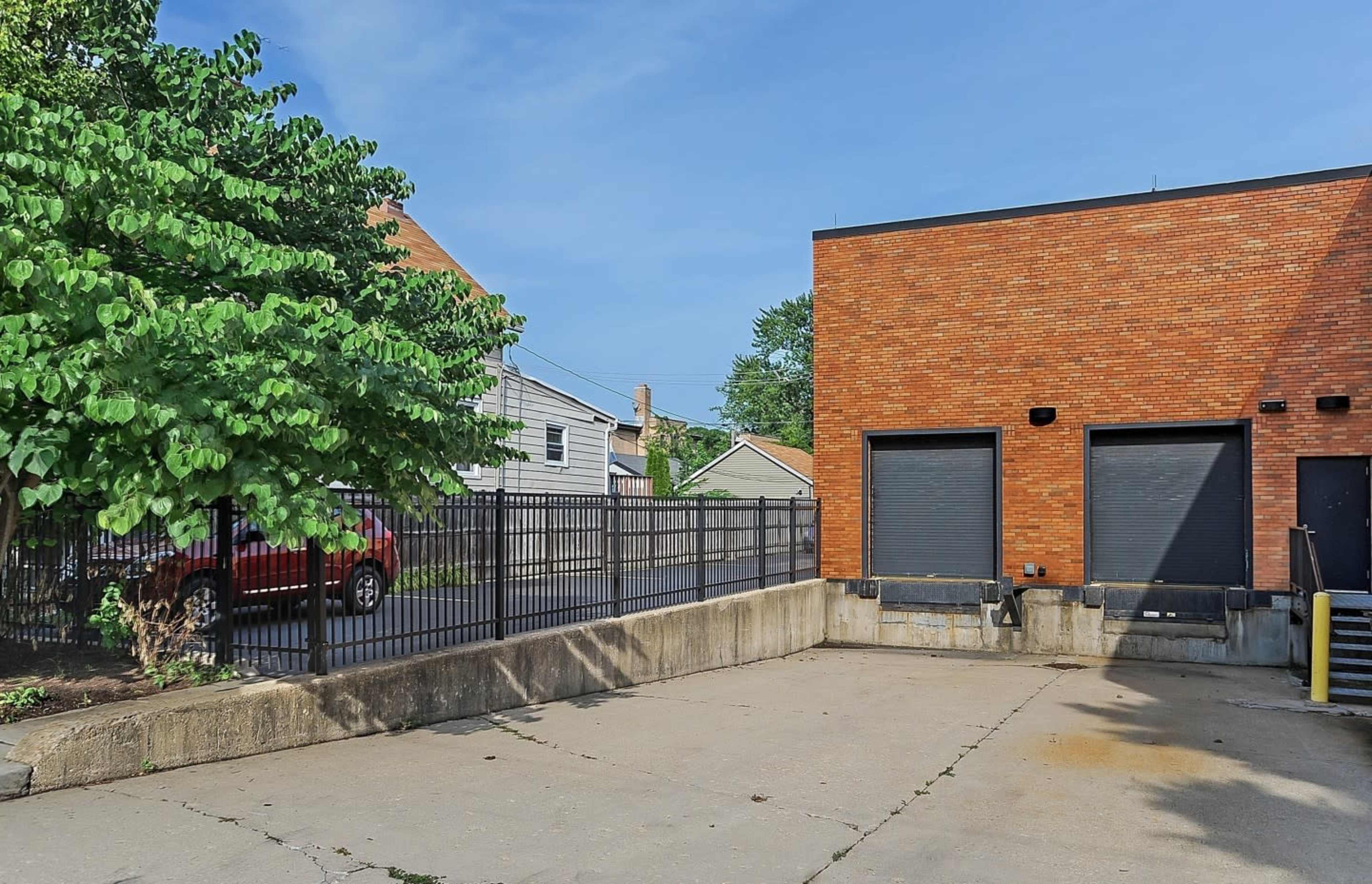 A brick building with closed metal doors is adjacent to a fenced concrete area, featuring a tree on one side and a parked red vehicle on a nearby street.