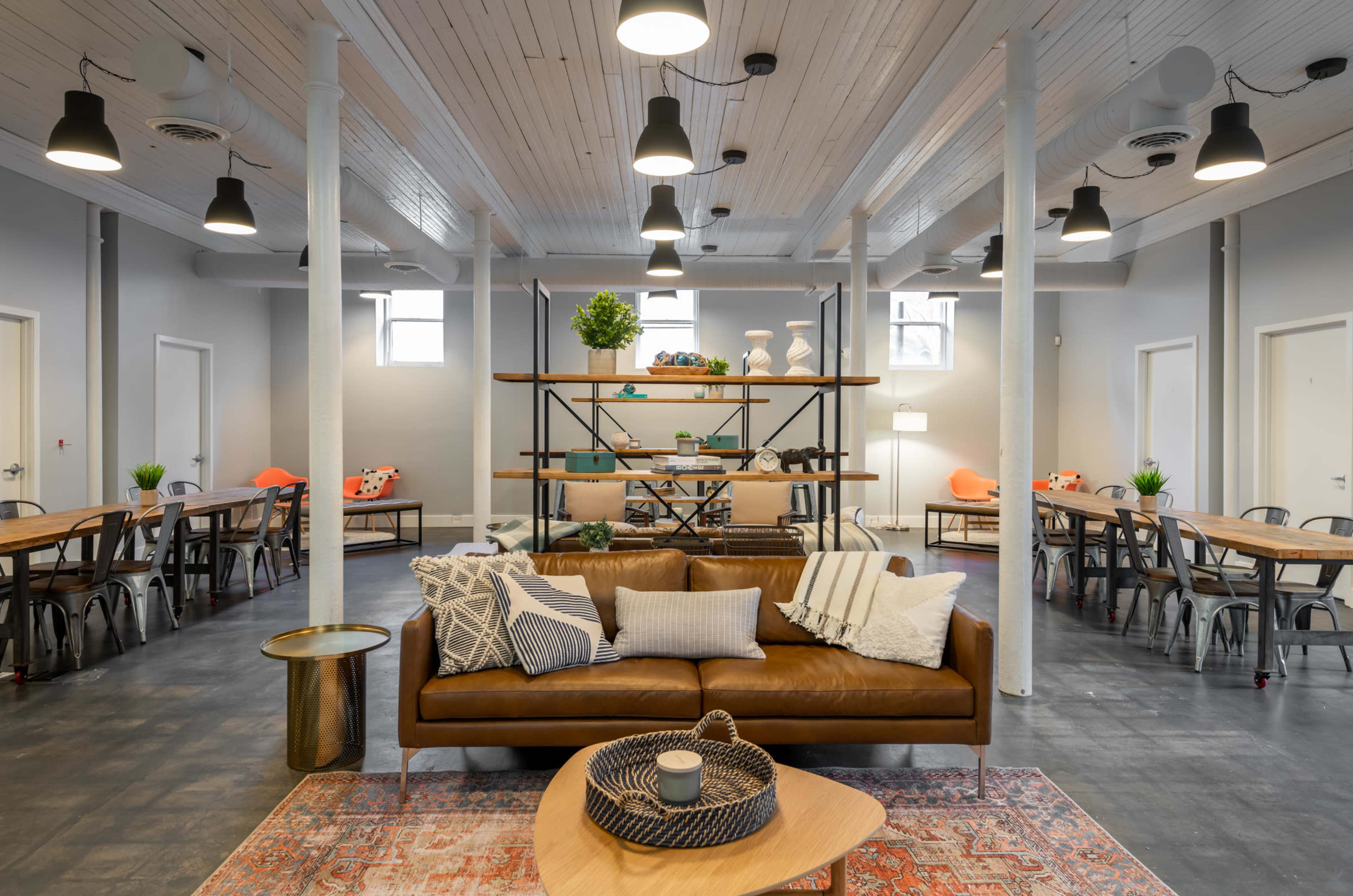 A modern, open communal space with a brown leather sofa, a wooden coffee table, and rows of tables and chairs arranged for dining or work.