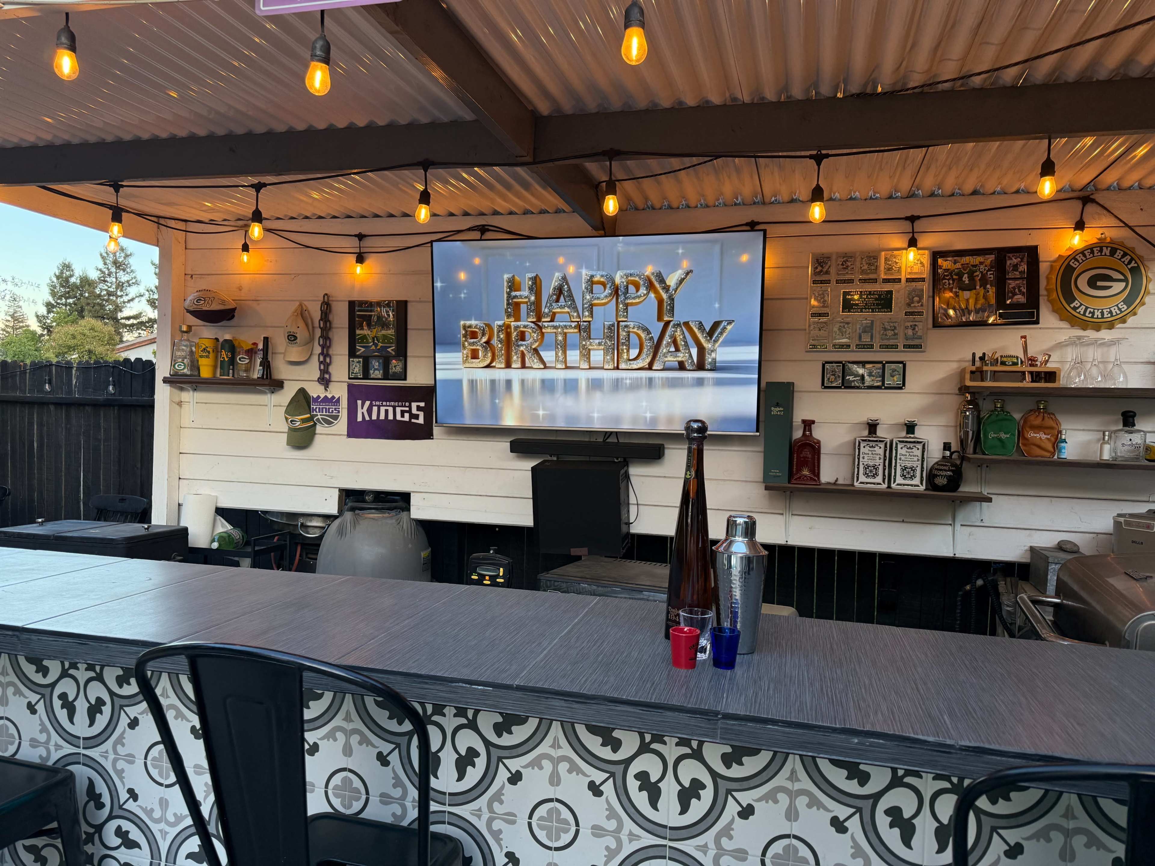 An outdoor bar area features a large television displaying a "Happy Birthday" message, with bottles and equipment arranged on the shelves behind it.