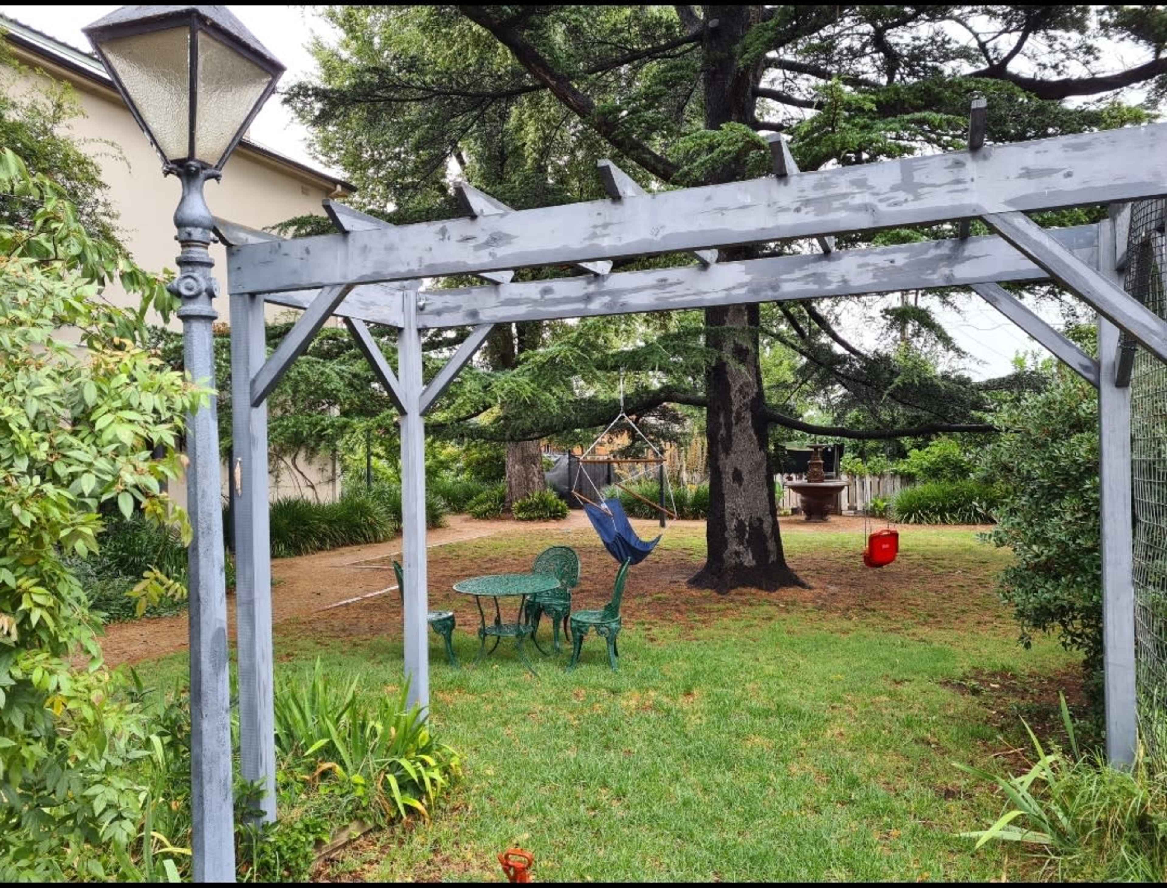 A wooden pergola with a lamp post frames a small seating area featuring a green table and chairs in a grassy garden.