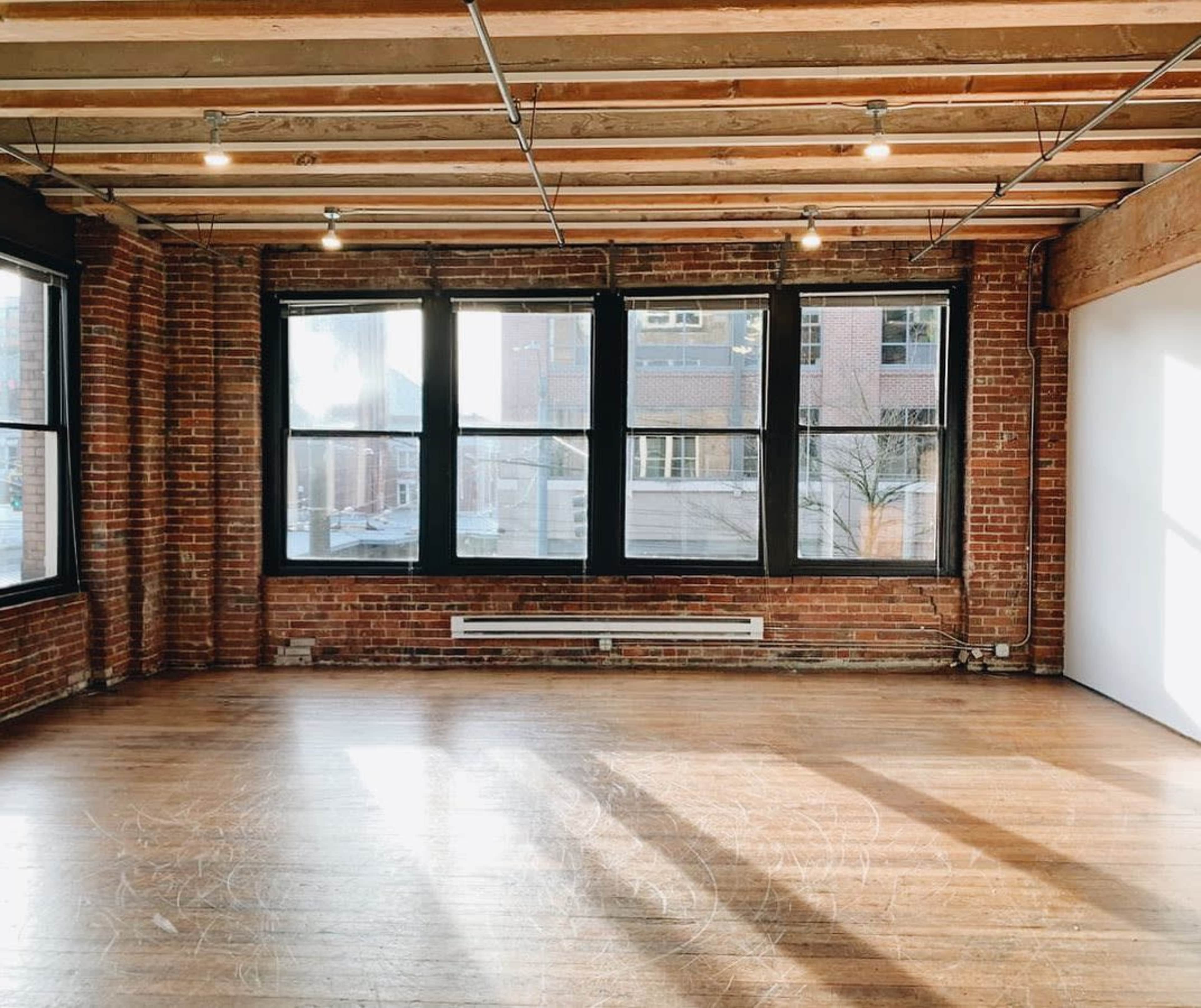 The image shows an empty room with exposed brick walls, wooden beams, and large windows allowing natural light to enter.