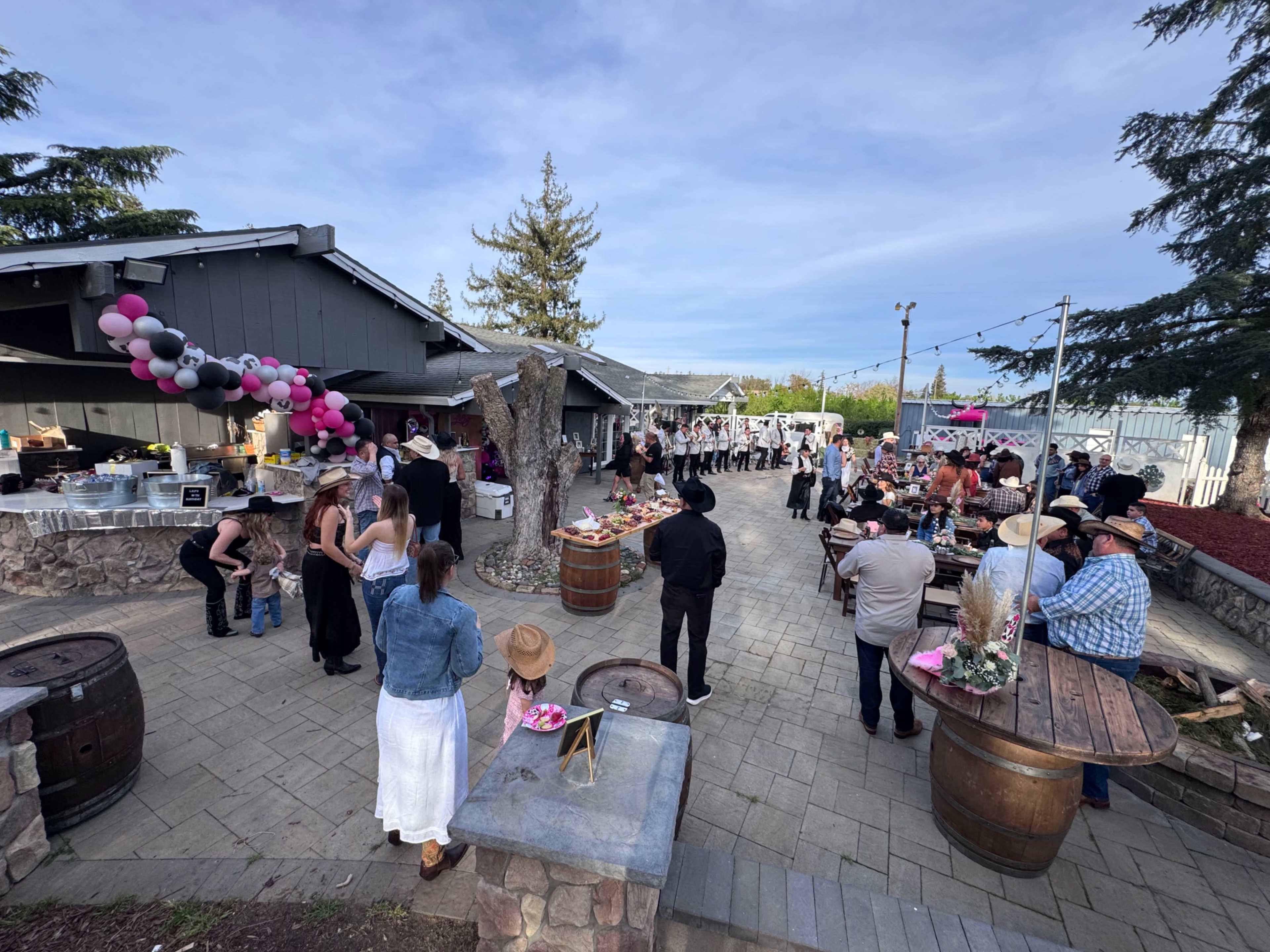 A gathering is taking place outdoors with guests mingling around a decorated courtyard featuring tables, barrels, and festive balloons.