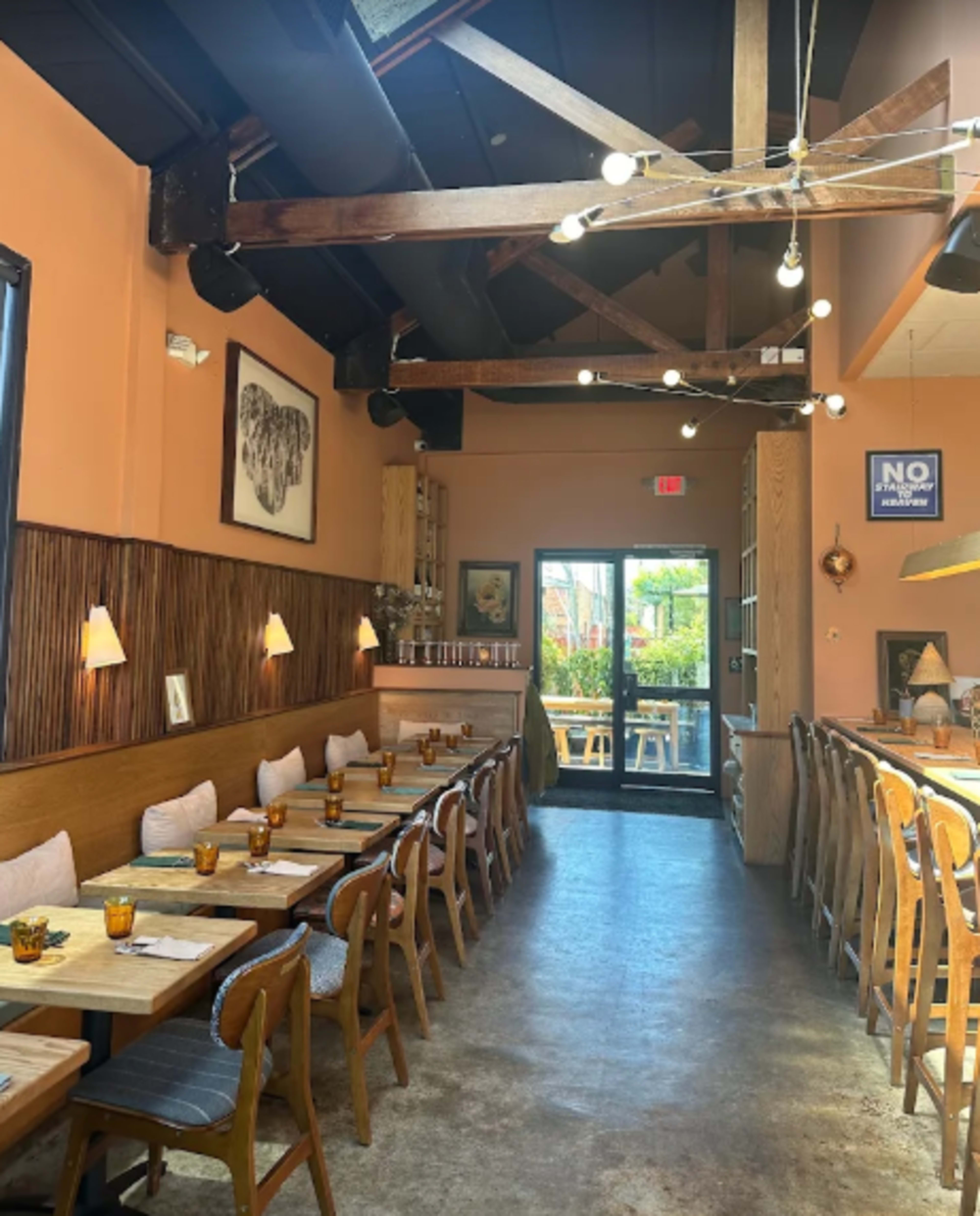 The image shows the interior of a restaurant with wooden beams, a long dining table lined with chairs, and light fixtures hanging from the ceiling.