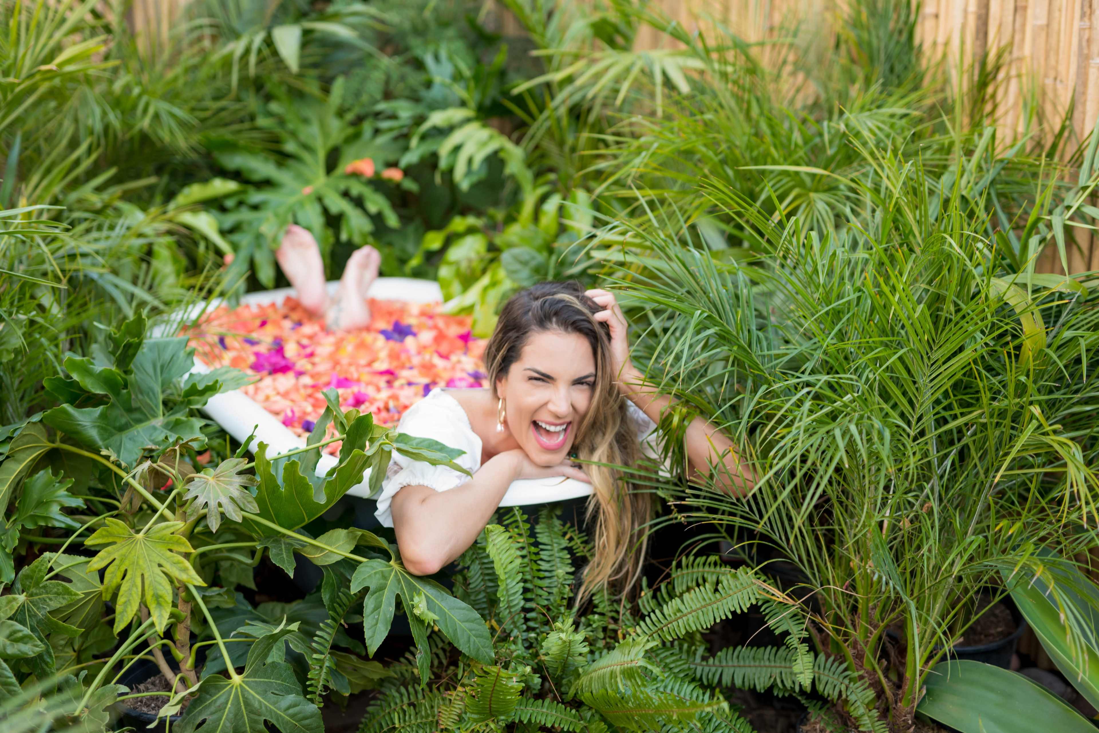 A woman is playfully lying in a bathtub surrounded by lush greenery and colorful flower petals.