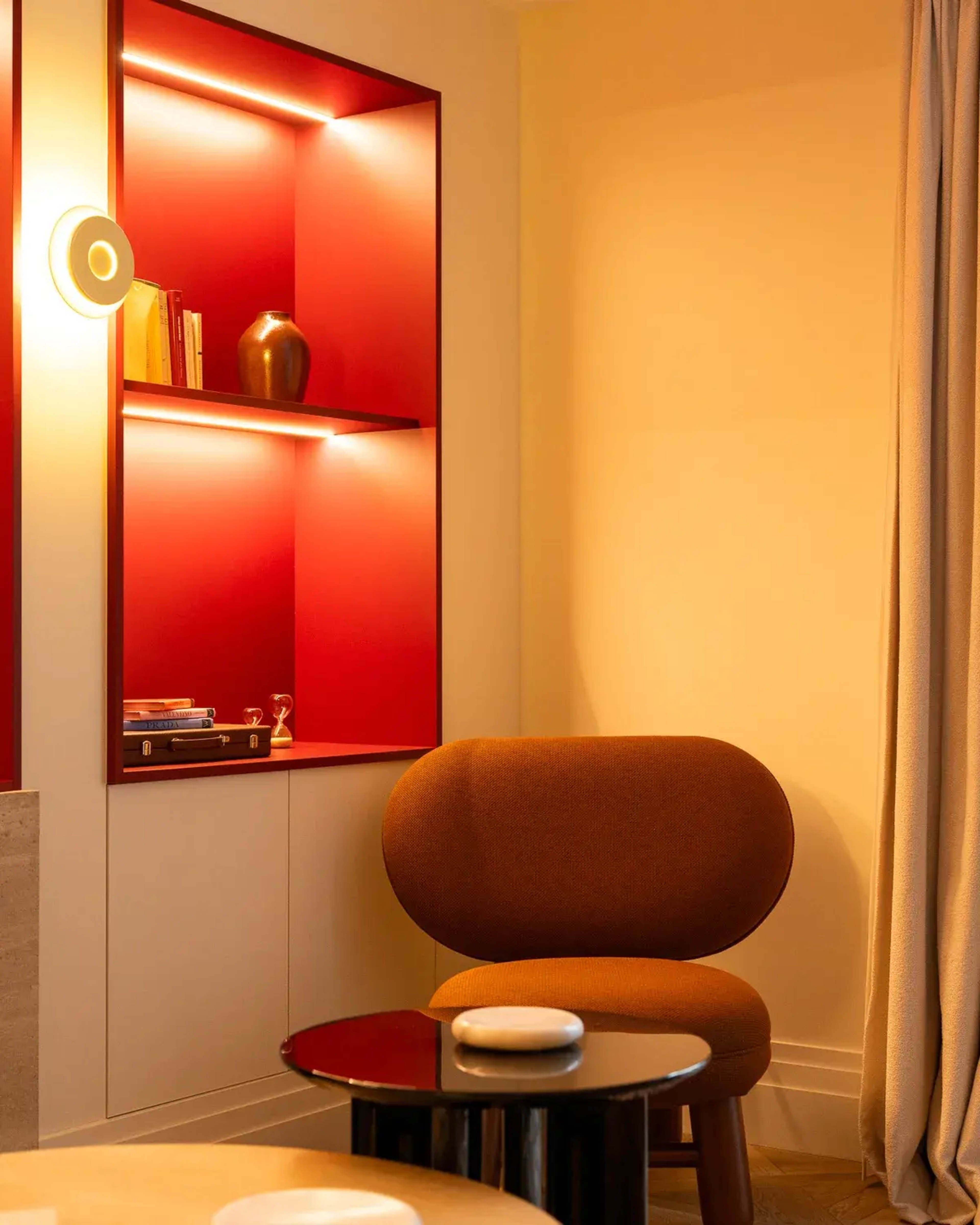 A cozy corner features a rounded brown chair next to a black table, with a shelf displaying books and a vase against a red backdrop.