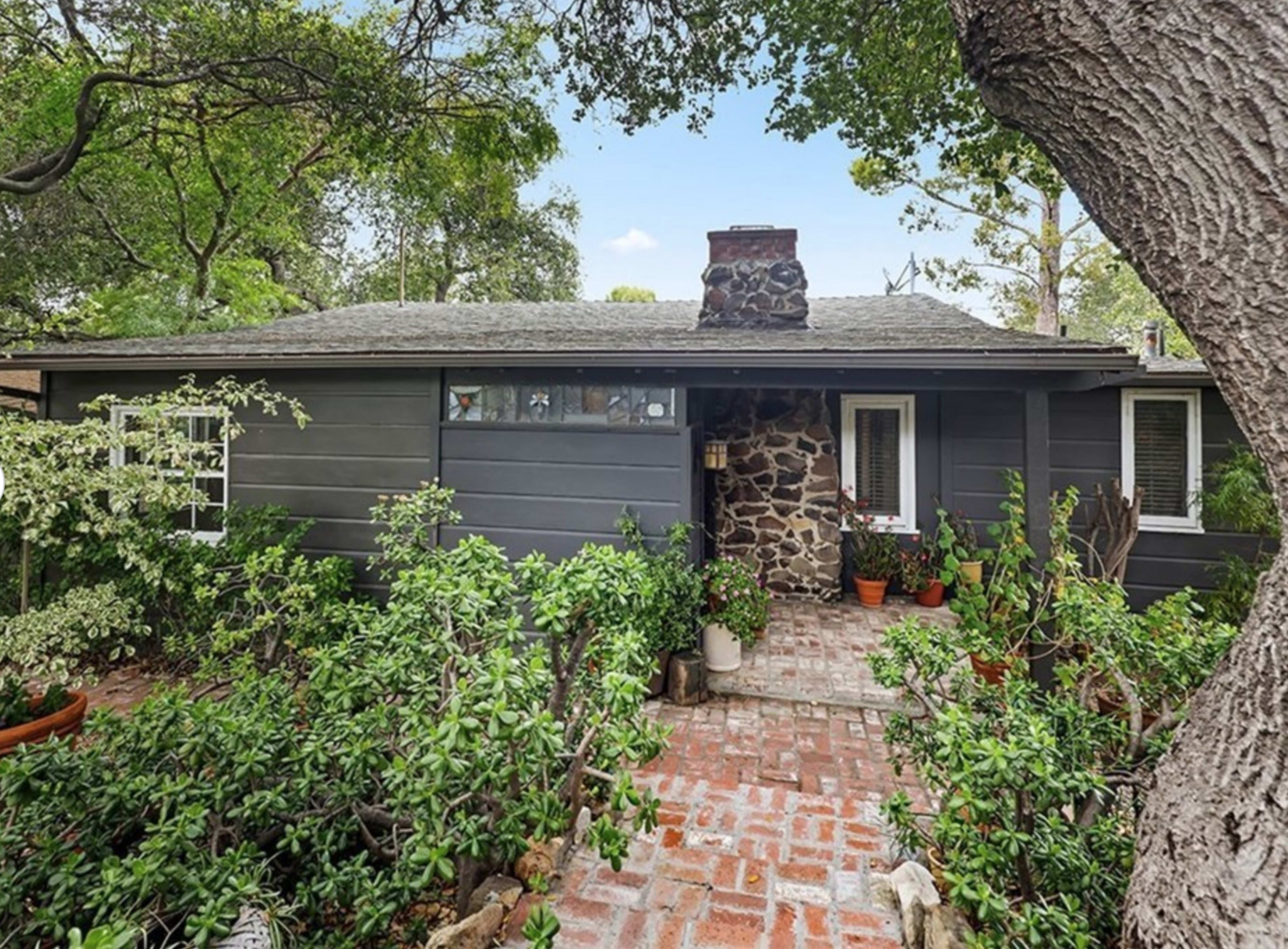 A single-story house with a stone chimney is surrounded by greenery and a brick pathway leading to the entrance.
