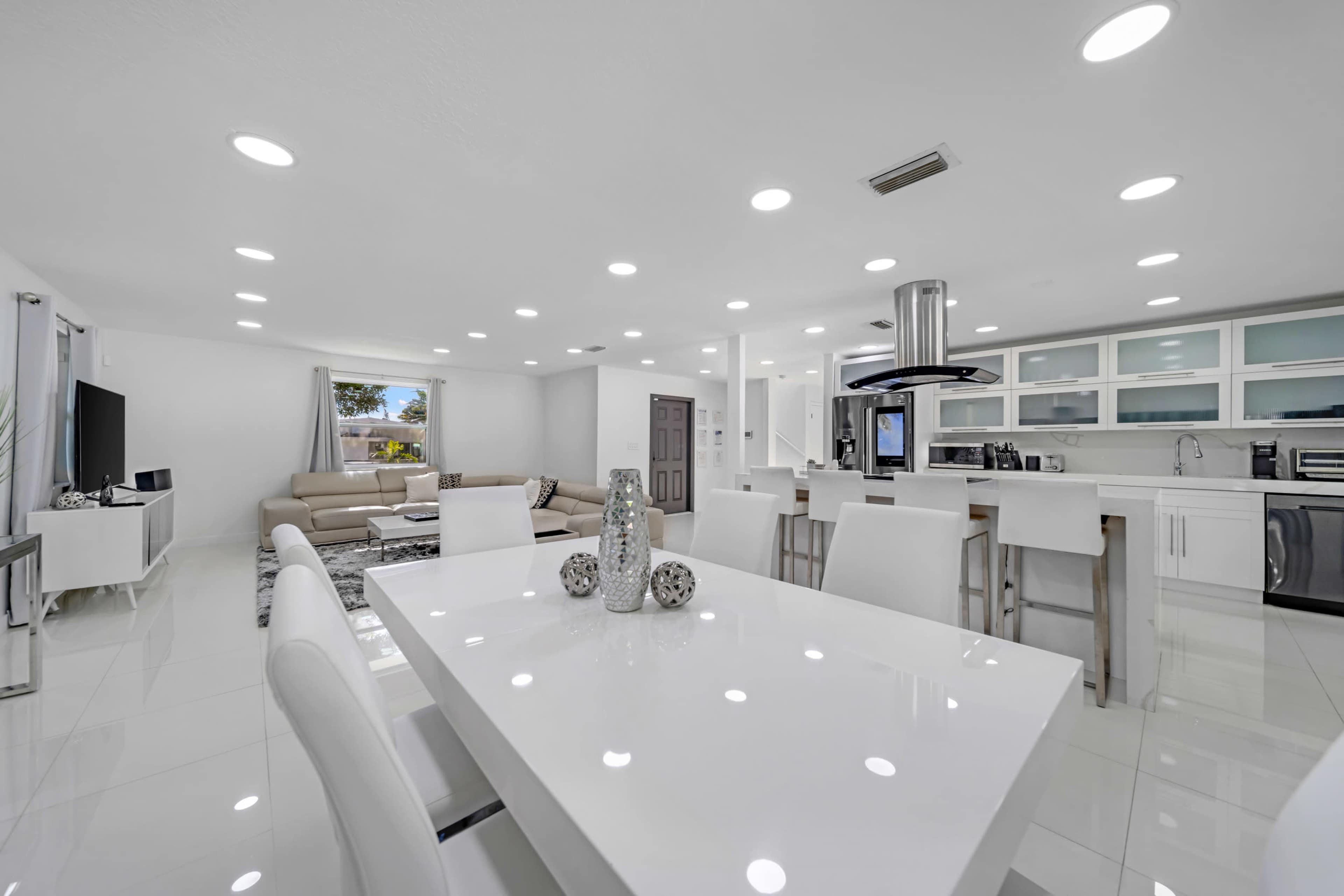 The image shows a modern, open-concept living and dining area with a sleek white table, comfortable seating, and a well-equipped kitchen.