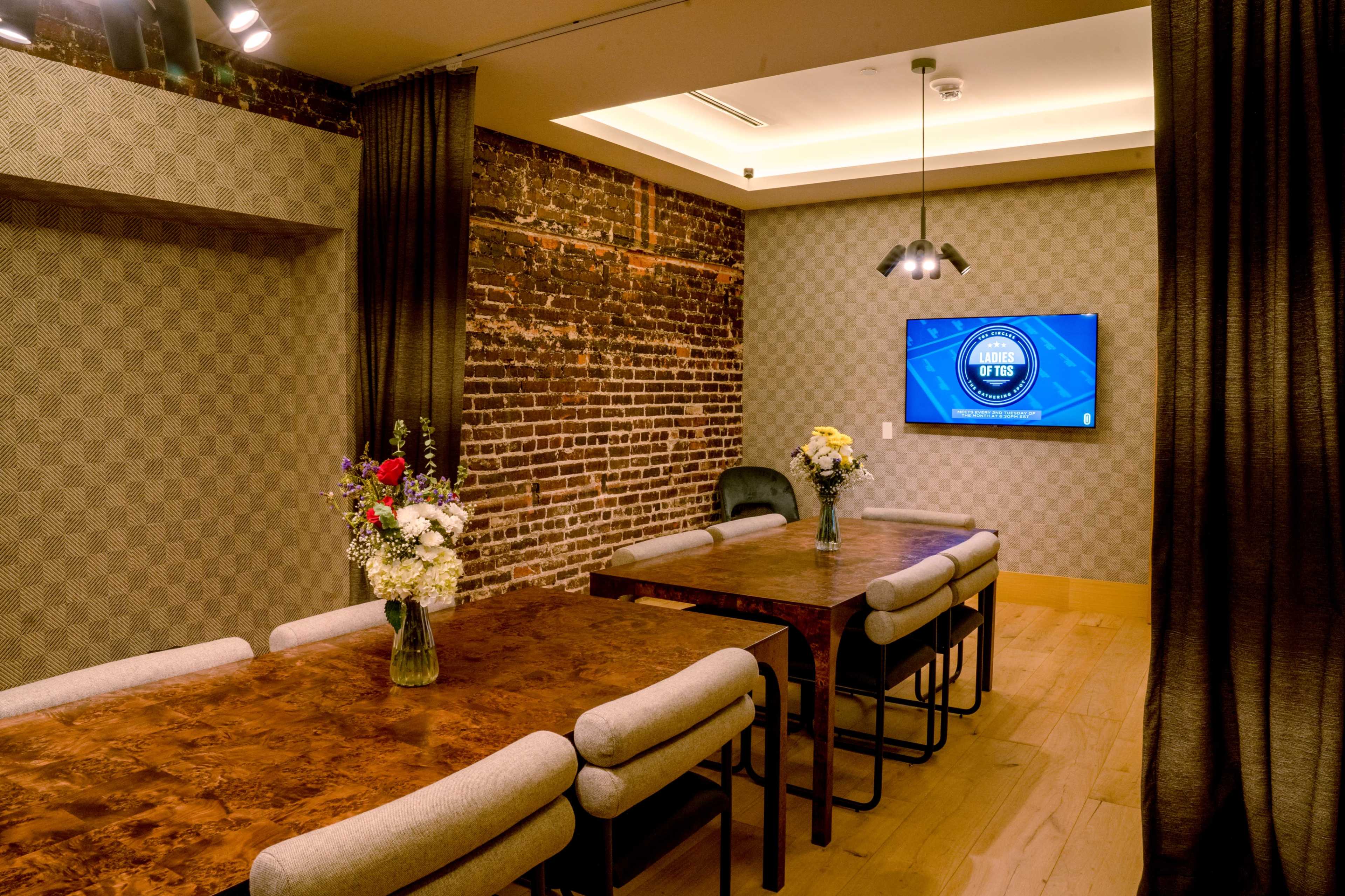 A conference room features a long wooden table surrounded by padded chairs, with a large screen mounted on the wall and floral centerpieces on the table.