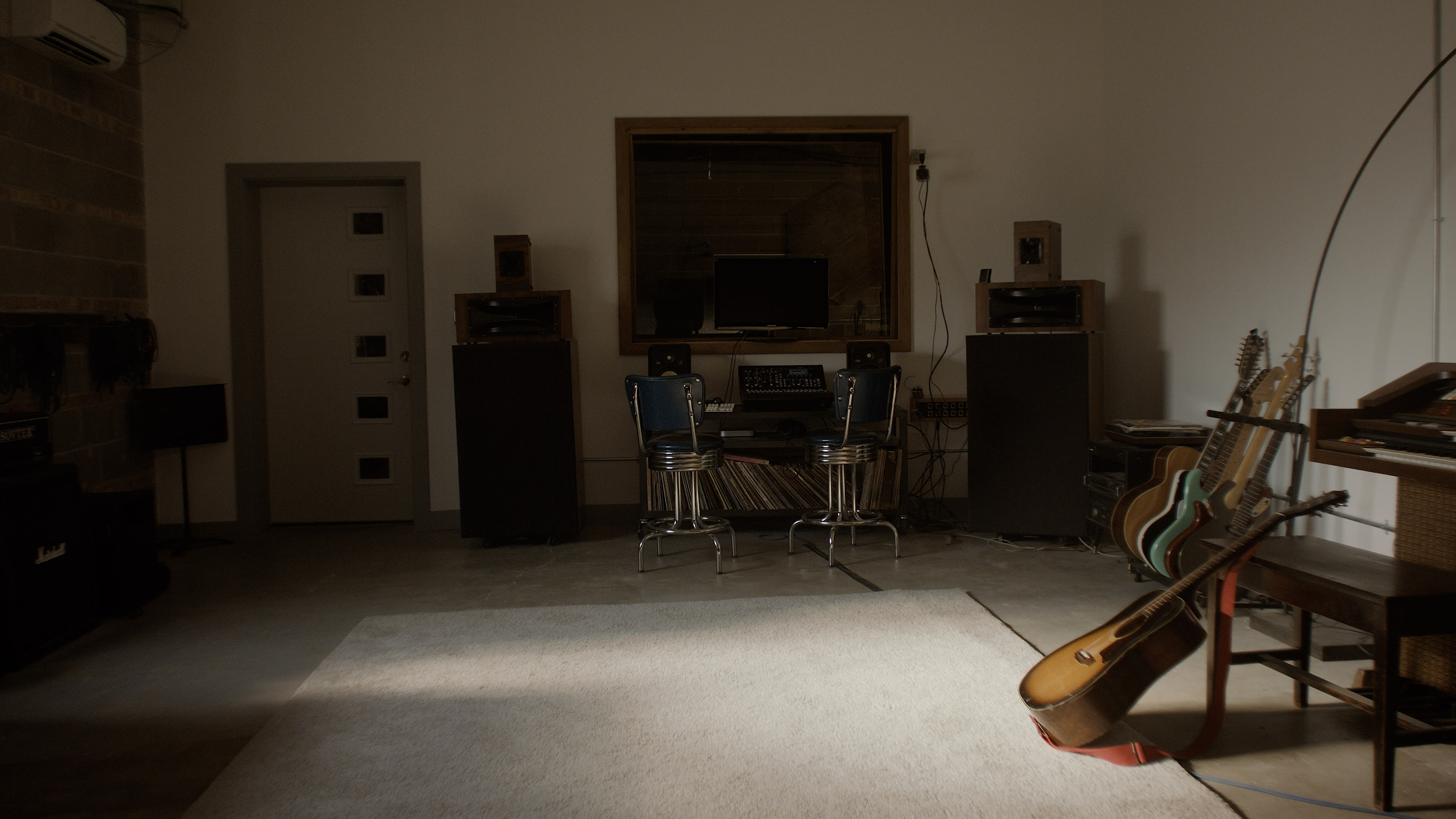 The image shows a minimalist music room featuring two bar stools, a television set, and several musical instruments arranged around a large area rug.