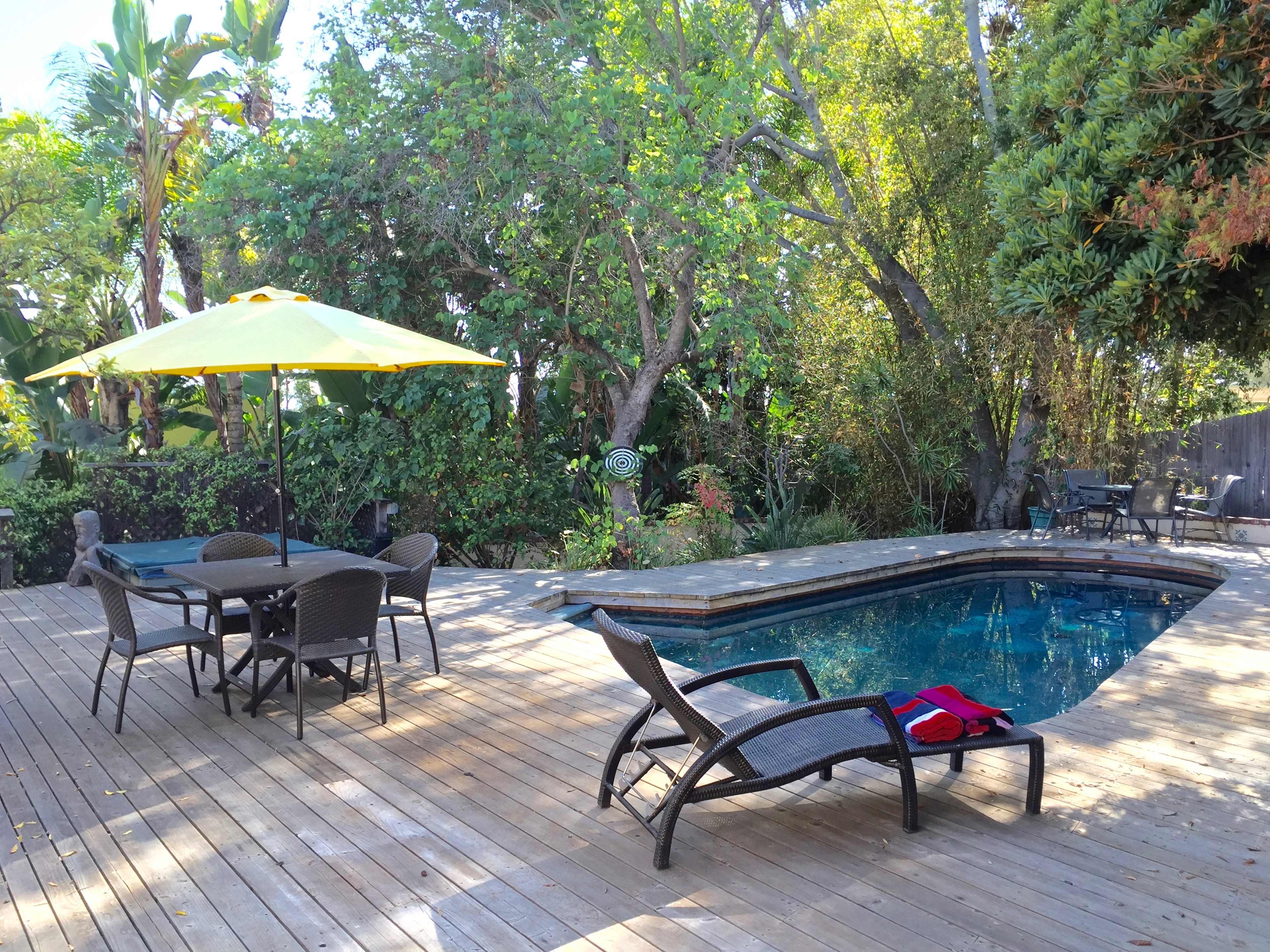 The image shows a backyard patio with a swimming pool, surrounded by lush greenery, featuring a table with chairs and a large umbrella.