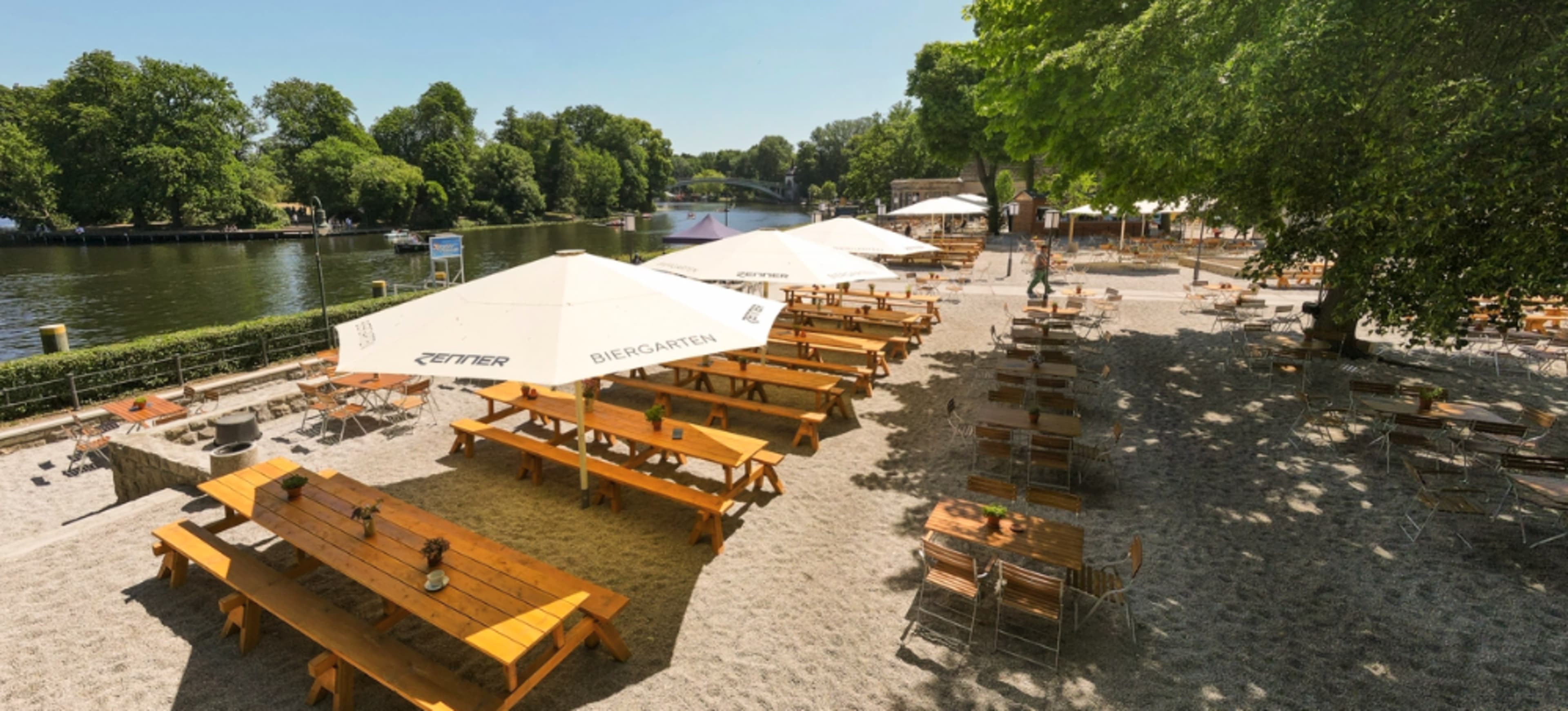 A riverside beer garden with wooden tables and chairs shaded by large umbrellas, surrounded by trees.