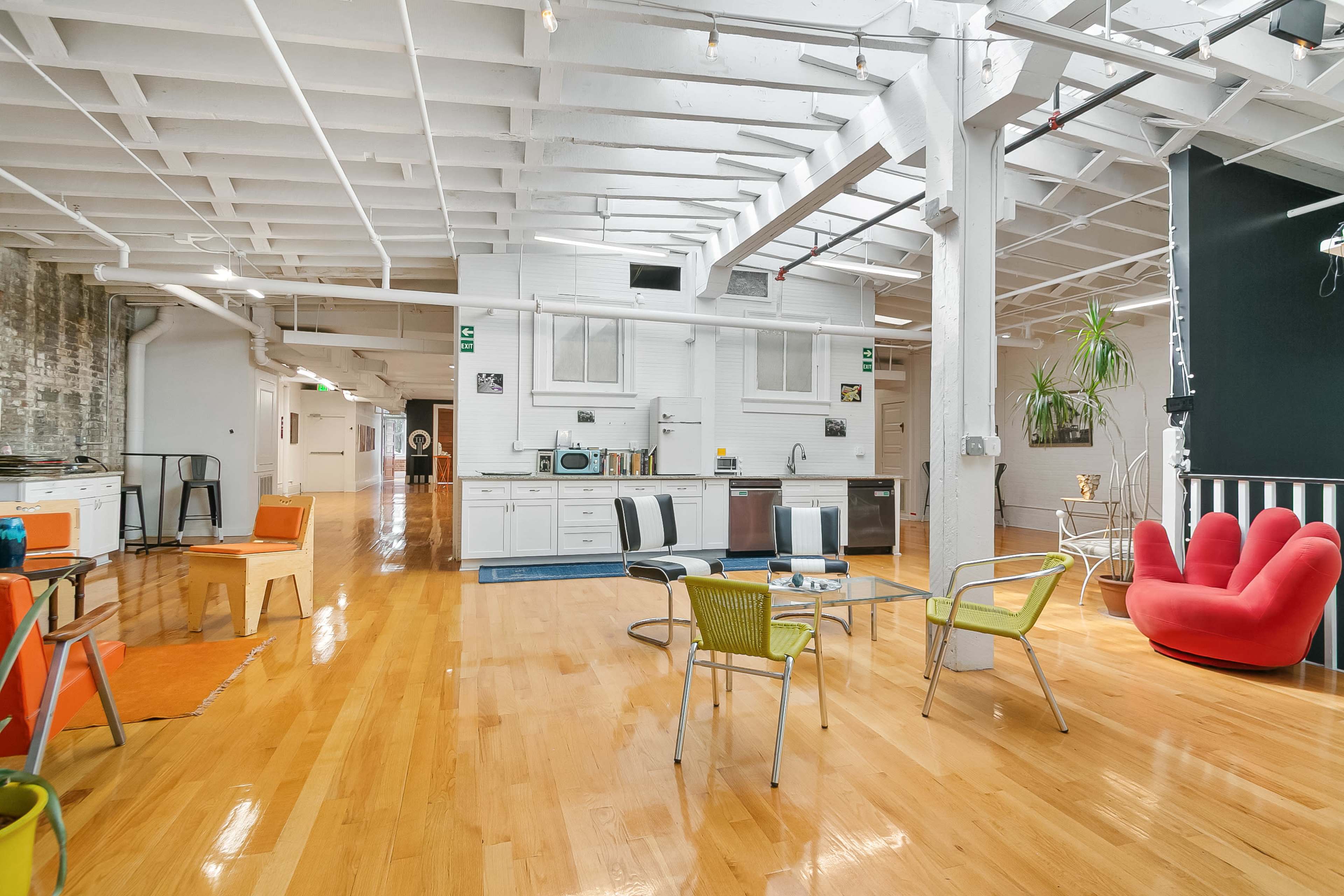 The image shows a spacious loft-style room with wooden floors, featuring a modern kitchen, colorful furniture, and large windows allowing natural light to enter.